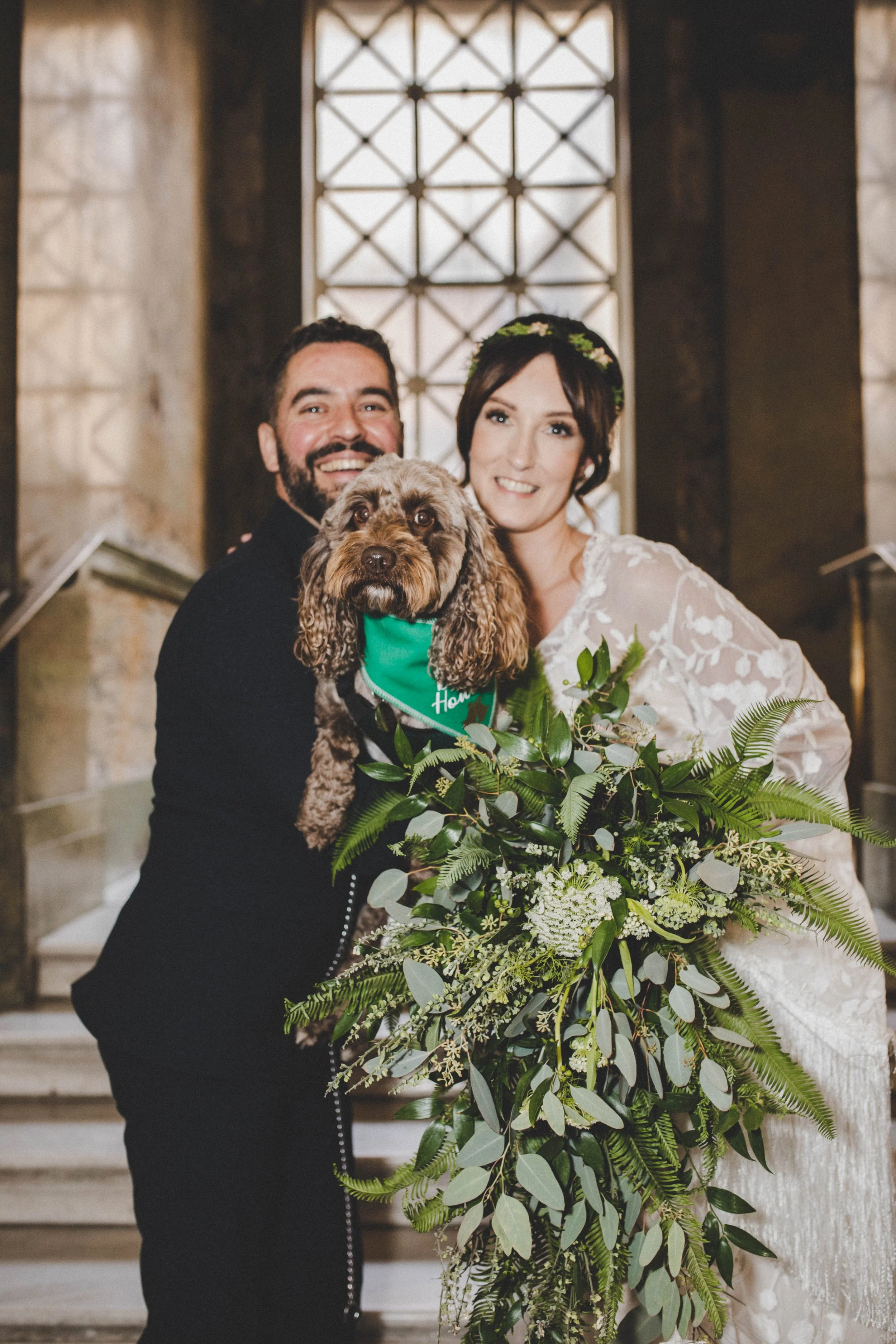A happy couple with a dog at a wedding, standing on stairs inside a church, with the bride holding a bouquet of flowers. The dog is held in front of them, wearing a green bandana.