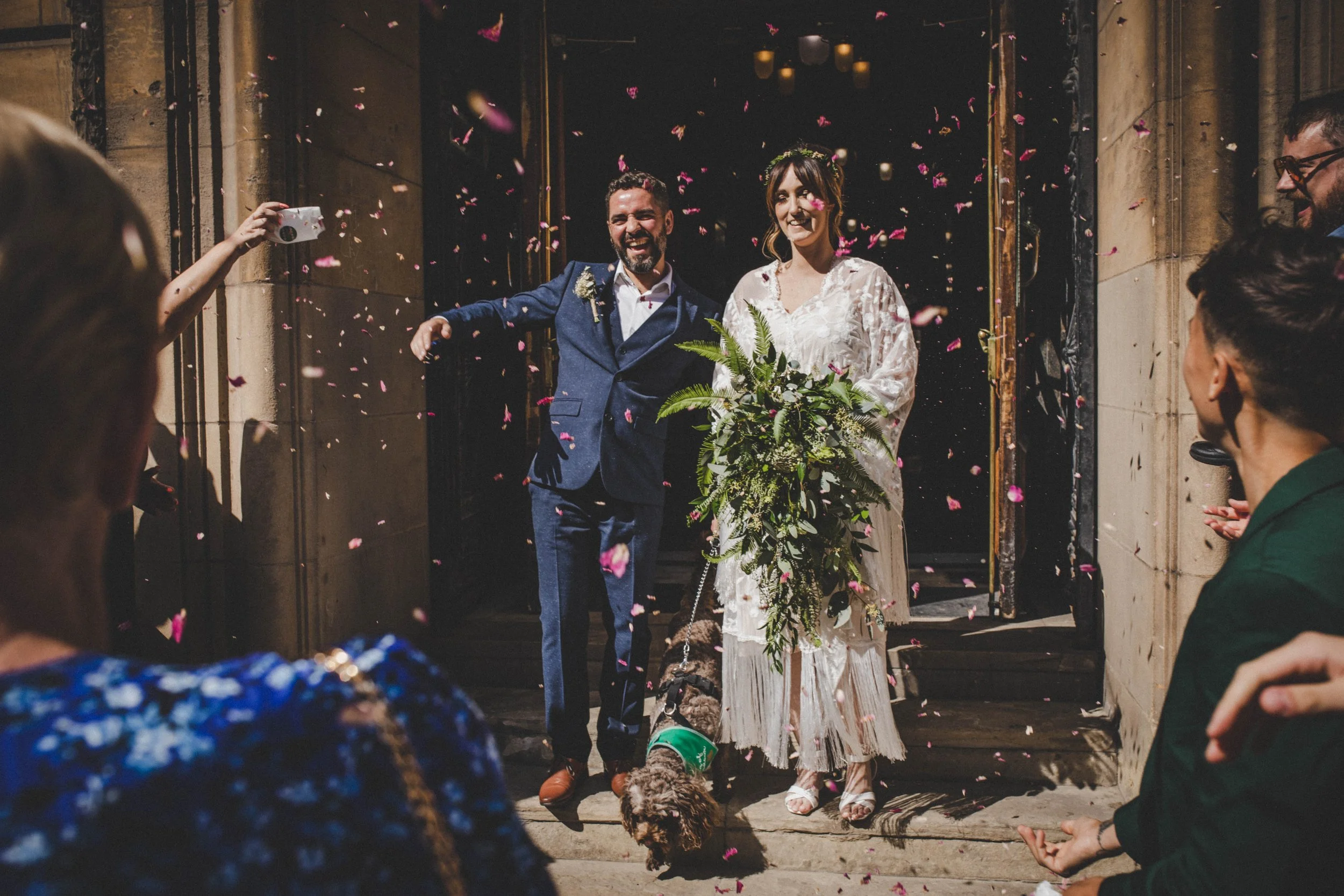 A bride and groom exiting a building, surrounded by guests throwing flower petals, celebrating their wedding.