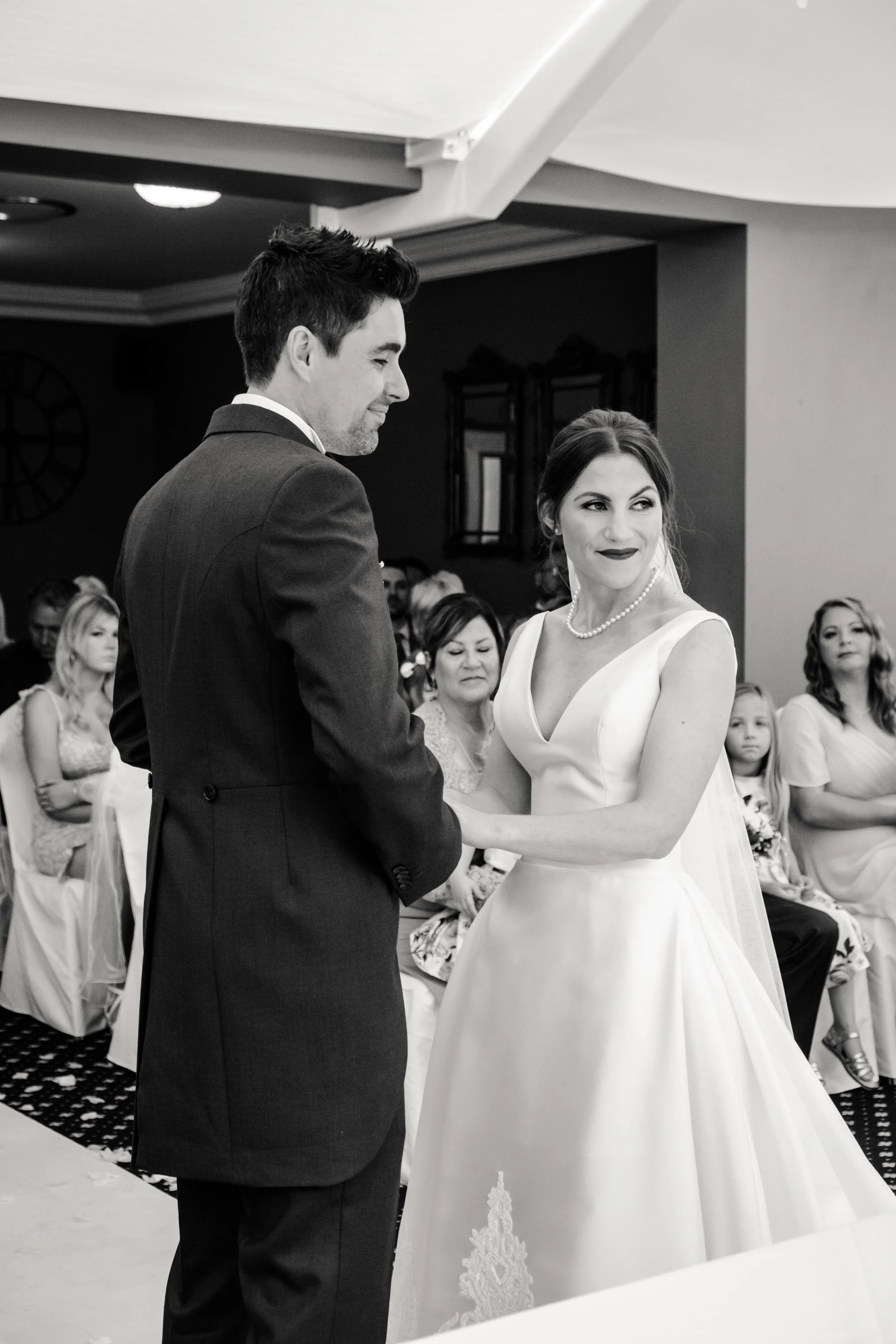 A woman and a man are holding hands during a wedding ceremony. The woman is wearing a white wedding dress and a pearl necklace, and the man is in a dark suit. They are smiling at each other with guests seated behind them.