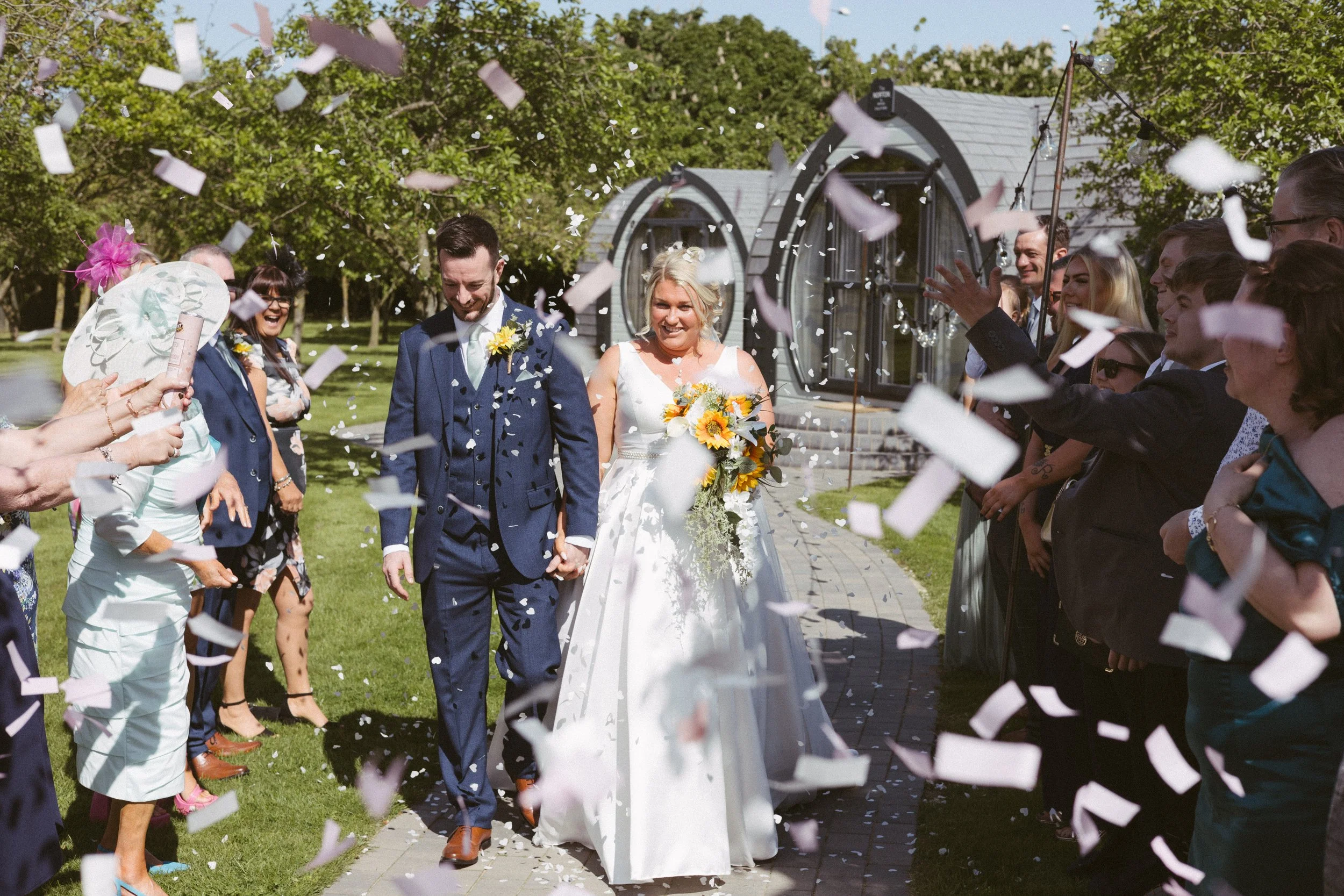 Bride and groom walking hand in hand through a confetti-throwing crowd on a sunny day during a wedding celebration.