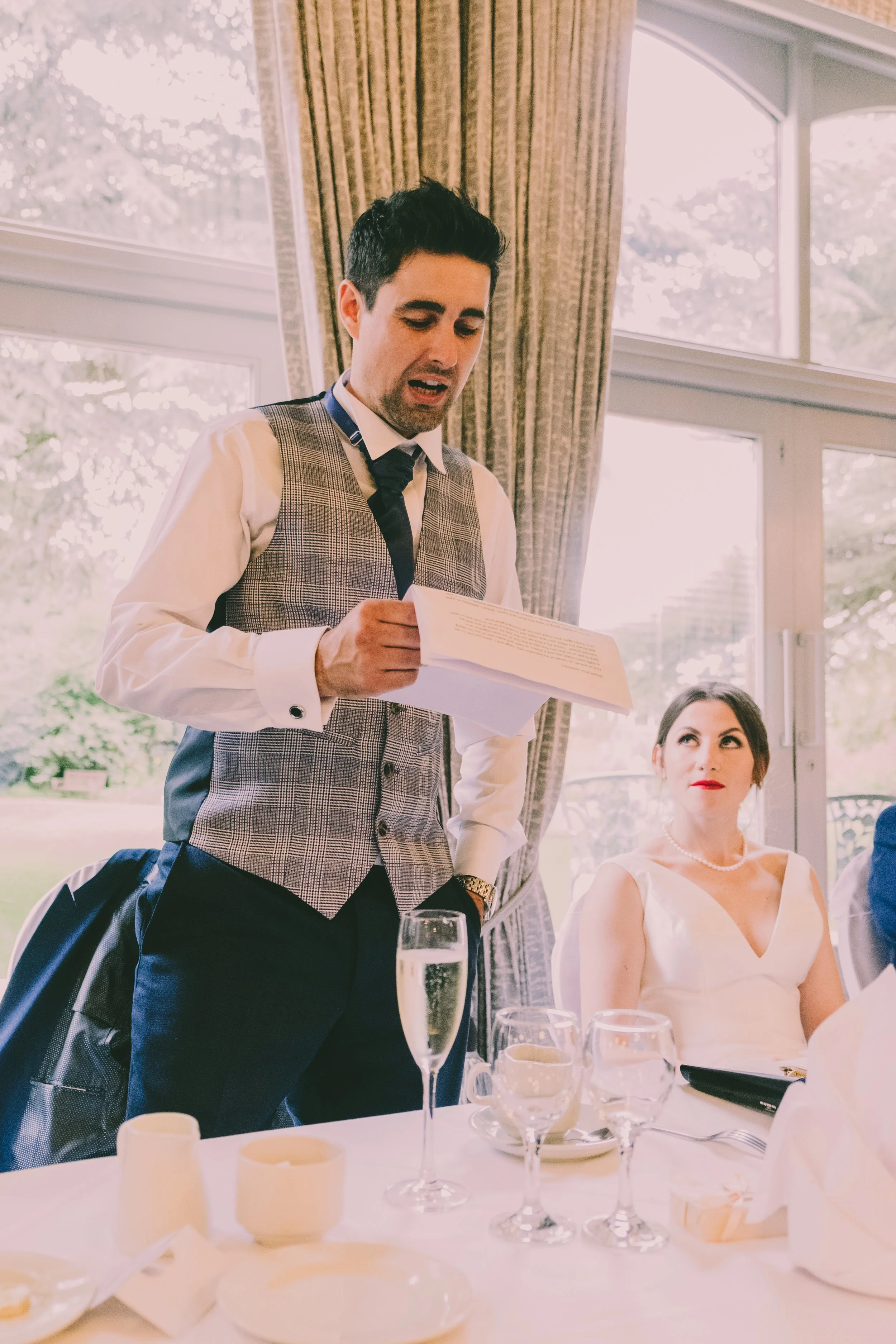 A man in a tuxedo with a plaid vest and a woman in a white dress sitting at a table during a formal event, with glassware and table settings, inside a bright room with large windows and curtains.