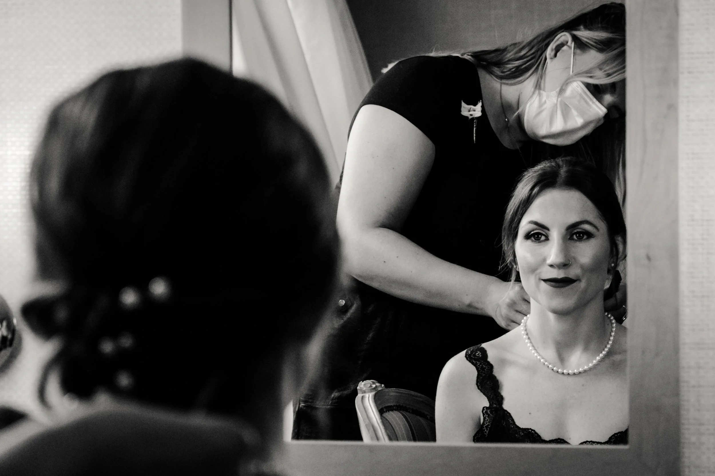 A woman with dark hair, wearing a pearl necklace and lace dress, sitting in front of a mirror with another woman applying makeup or making an adjustment behind her, both wearing masks.