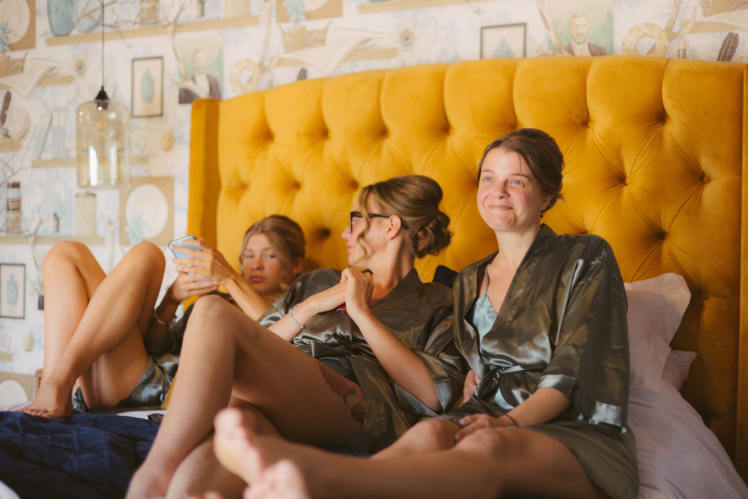 A group of friends sitting on a bed with a yellow headboard, in a cozy room, relaxing and smiling.