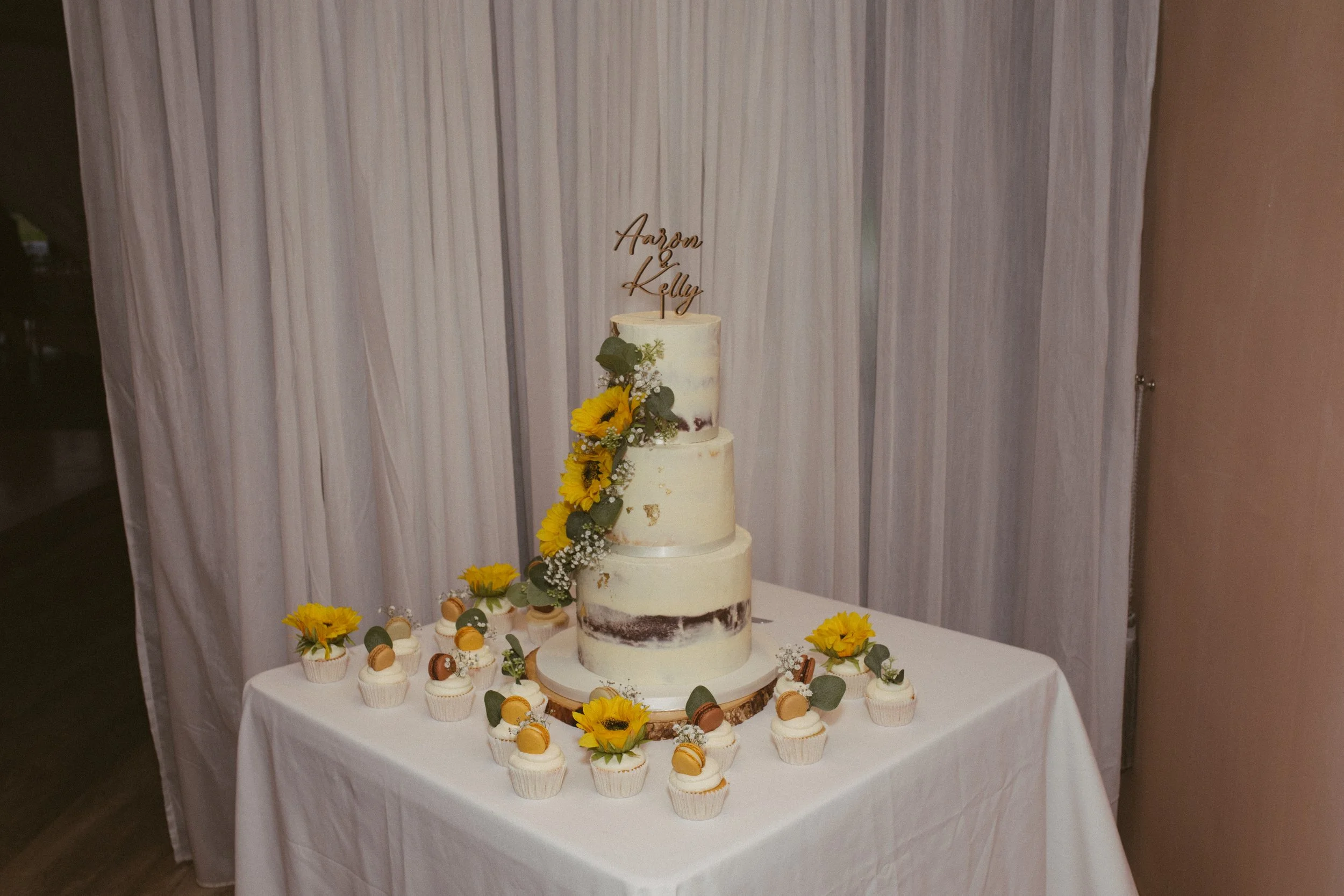A tiered wedding cake with sunflower and greenery decorations on a white table, surrounded by cupcakes decorated with macarons and flowers, with a backdrop of light-colored curtains.