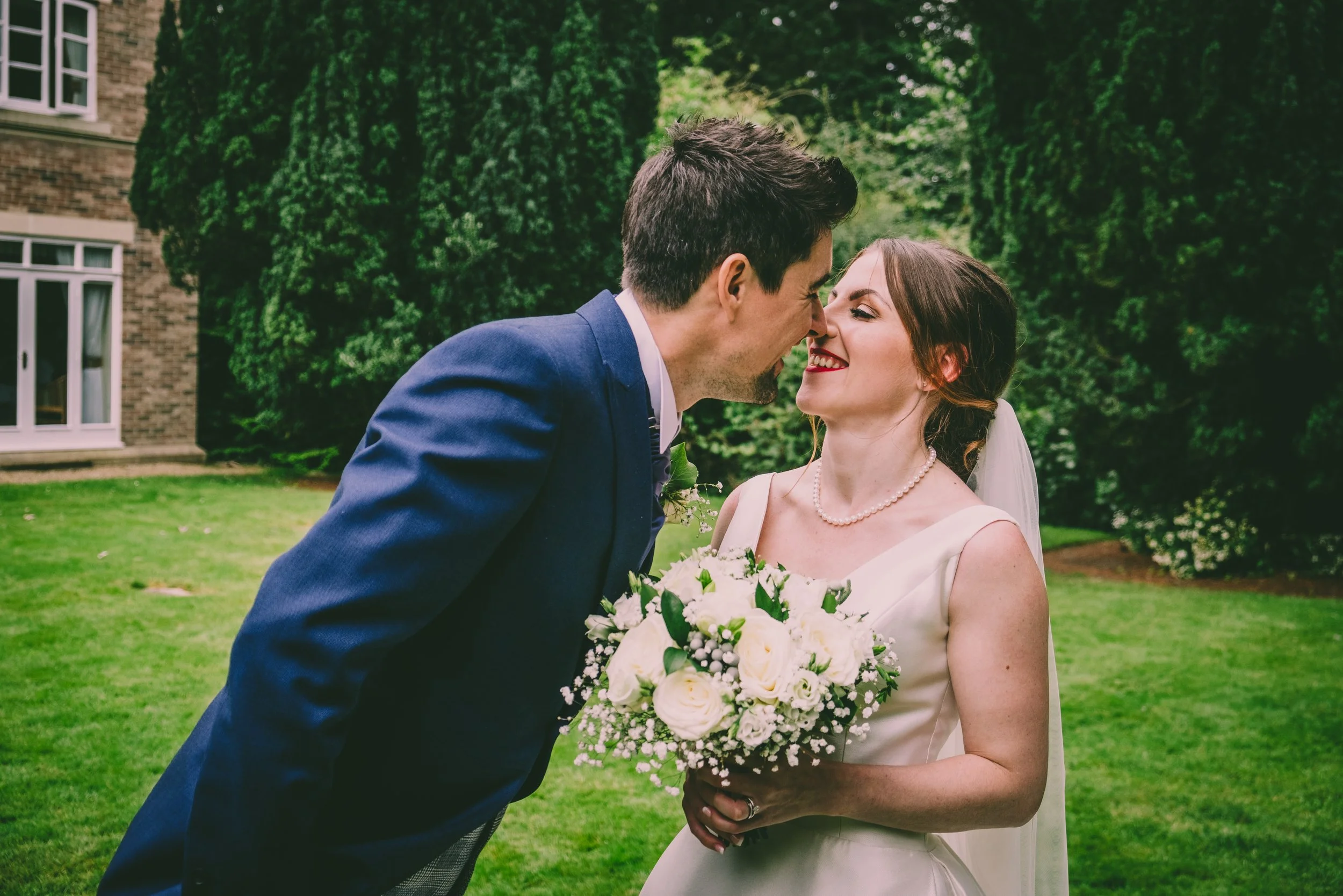 A bride and groom sharing a kiss outdoors during their wedding, with the bride holding a bouquet of white and pink flowers and wearing a pearl necklace, in front of green trees and a brick building.