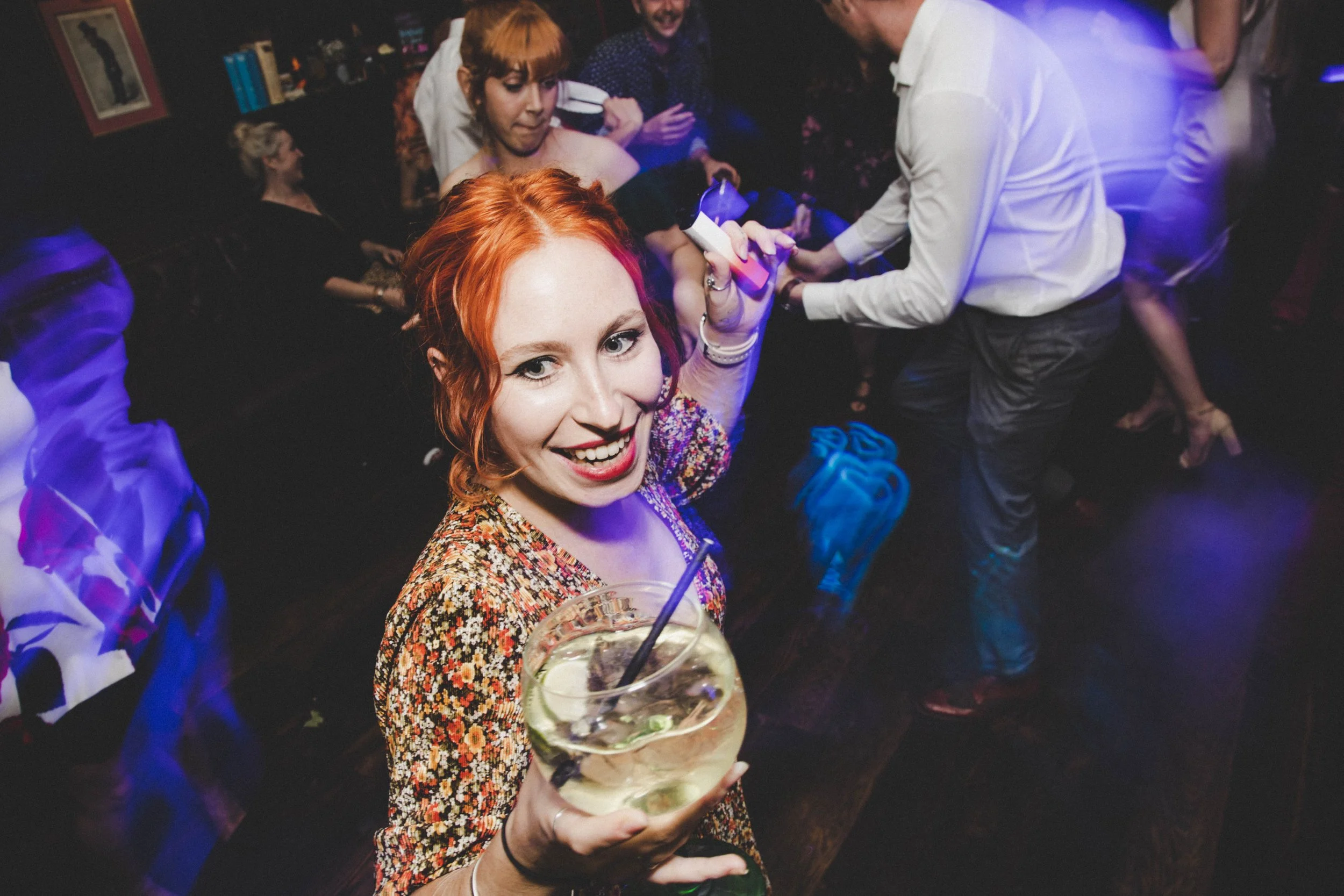 A woman with red hair smiling at the camera while holding a large cocktail in a bar or nightclub. Other people are in the background, socializing and enjoying drinks.