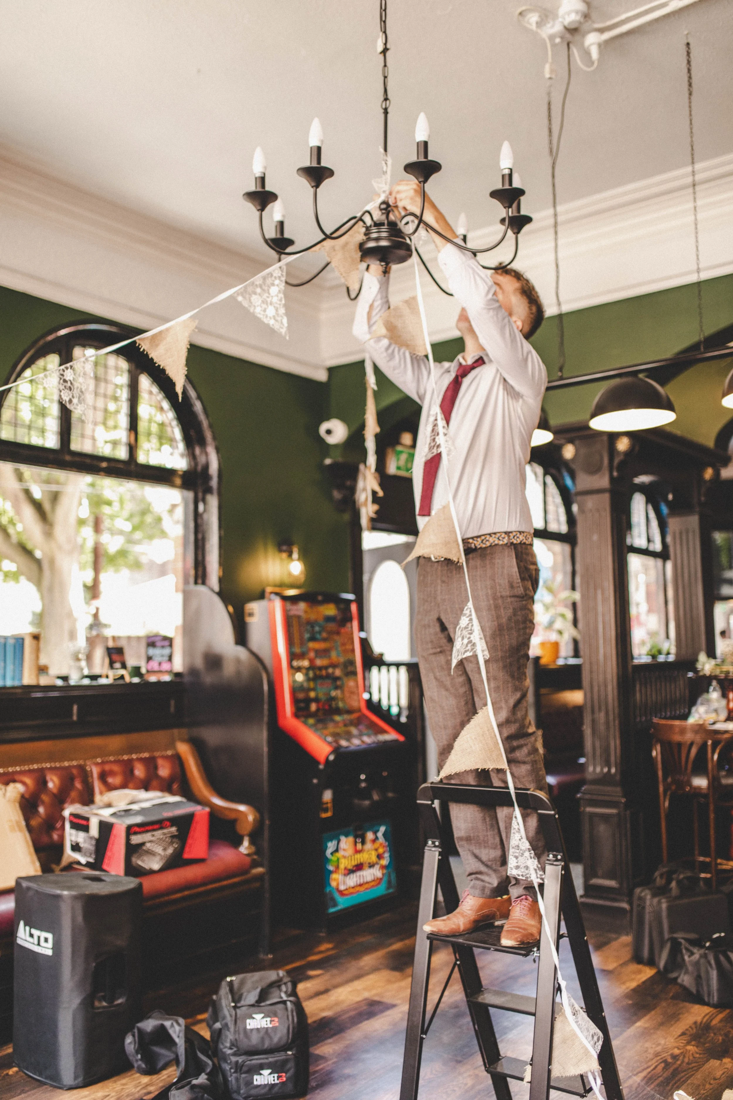 A man in a white shirt, red tie, and brown pants standing on a ladder, hanging decorative bunting in a room with dark wooden floors, green walls, and large arched windows, with arcade games and a piano in the background.