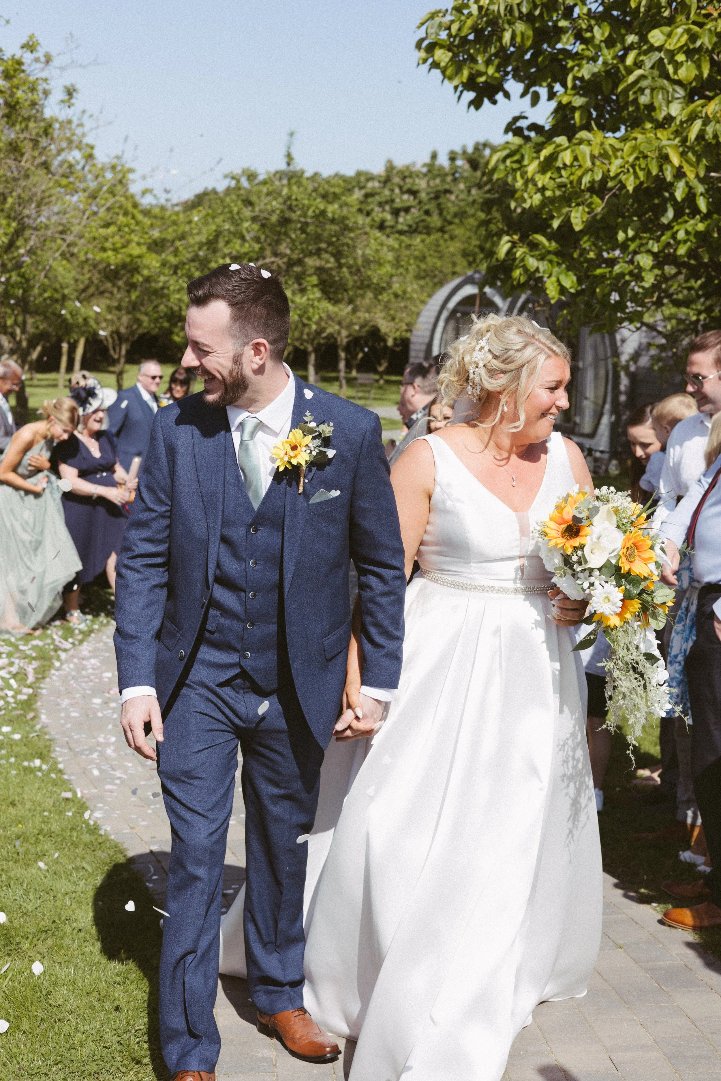 A bride and groom holding hands at an outdoor wedding ceremony, surrounded by guests, with greenery and trees in the background.