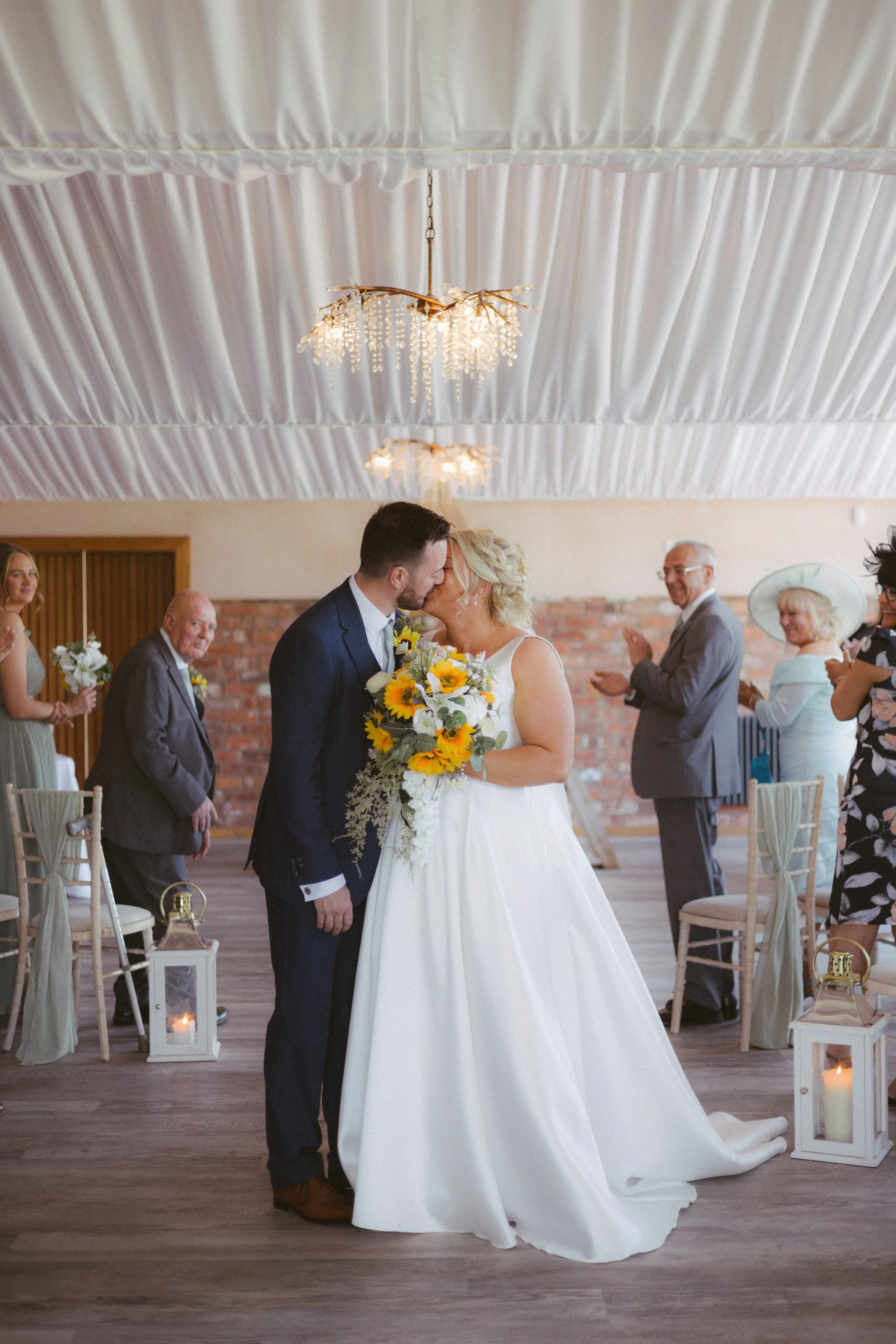 A bride and groom share a kiss during their wedding ceremony, surrounded by guests in a decorated indoor venue with white drapes, chandeliers, candles, and chairs.