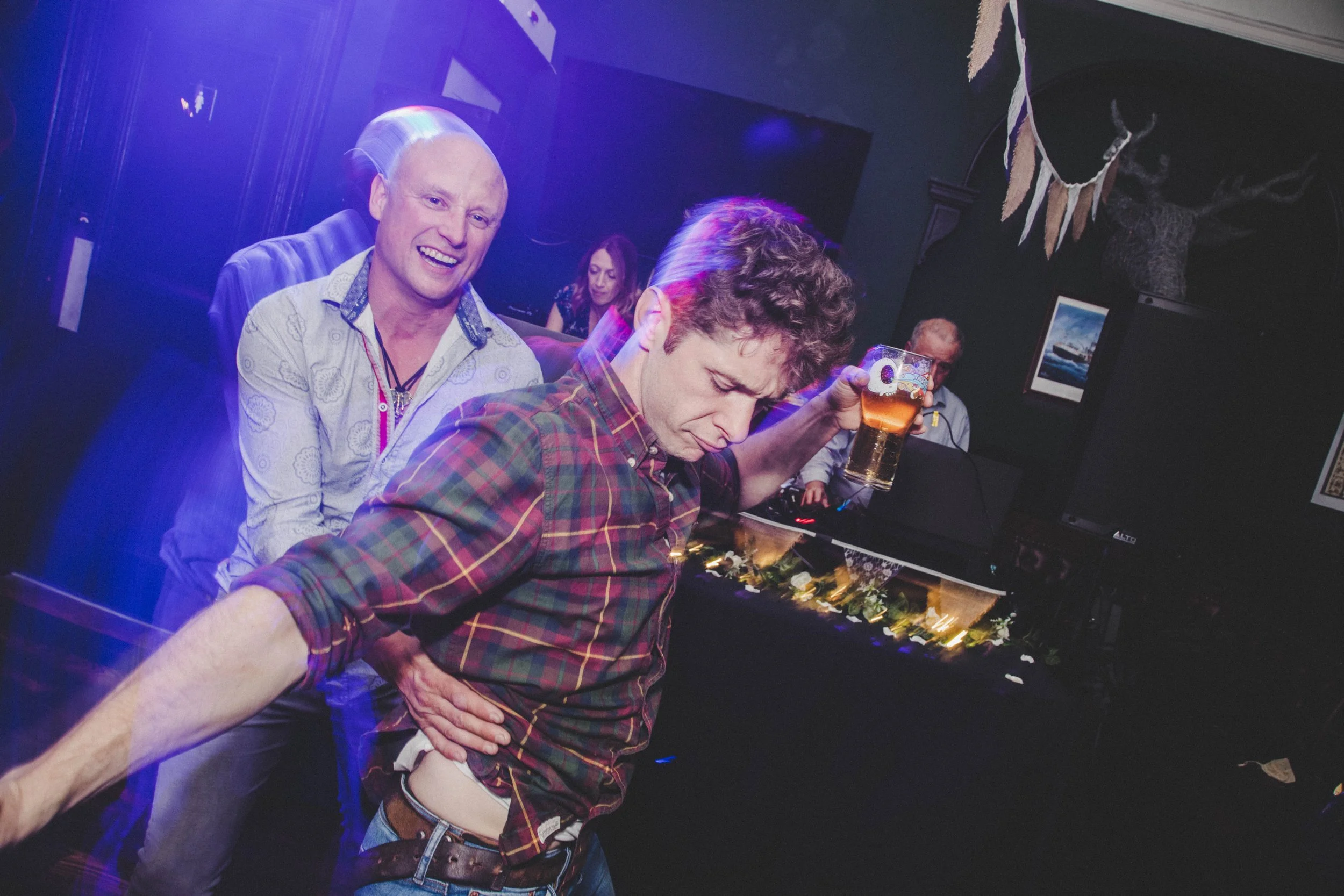 A group of people at a party or social gathering, with two men in the foreground. One man with short blonde hair and a patterned shirt is smiling, while another man with curly hair in a plaid shirt is holding a drink and looking down. In the backgrou
