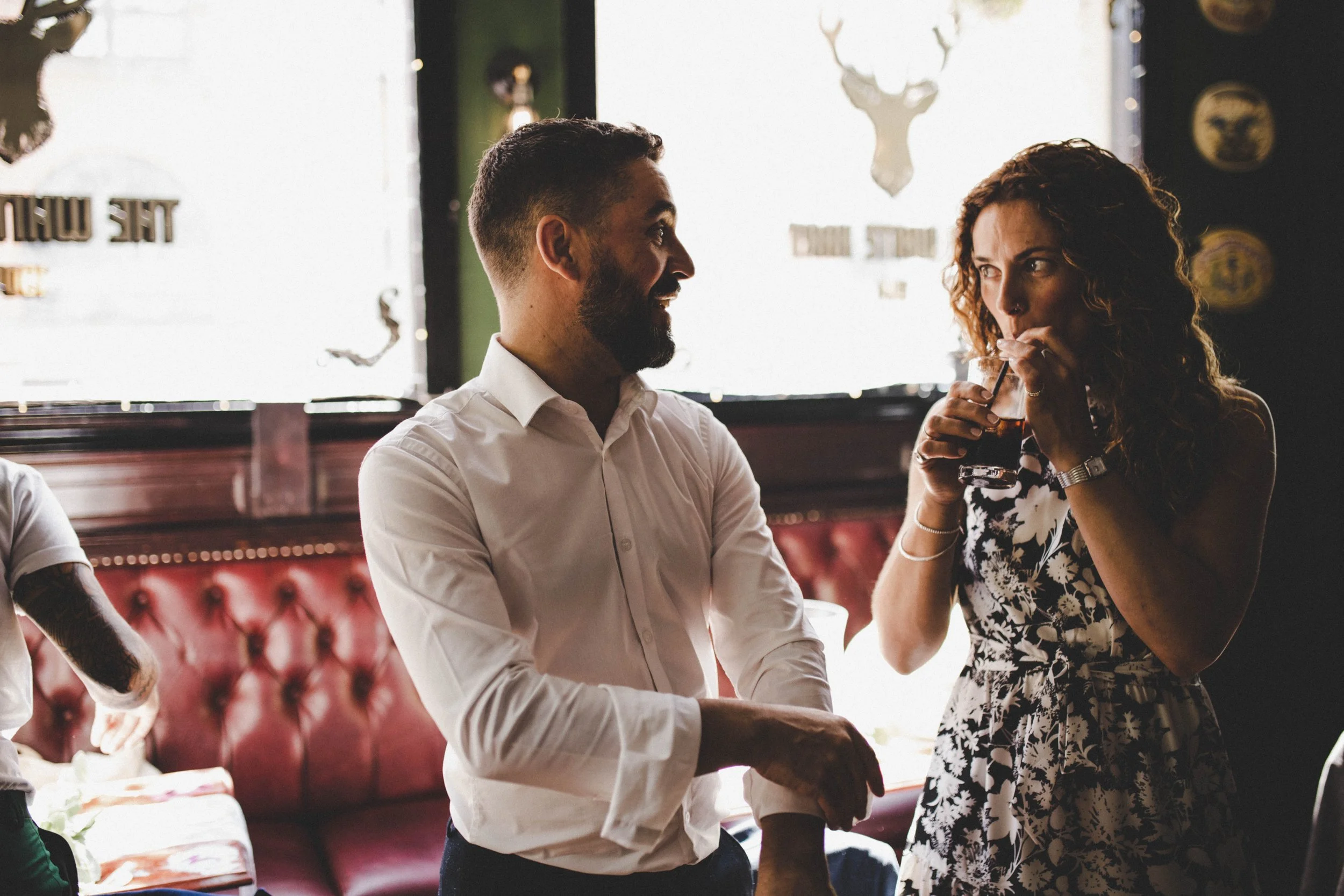 A man and woman enjoying a drink and conversation in a dimly lit restaurant or bar with a window in the background.