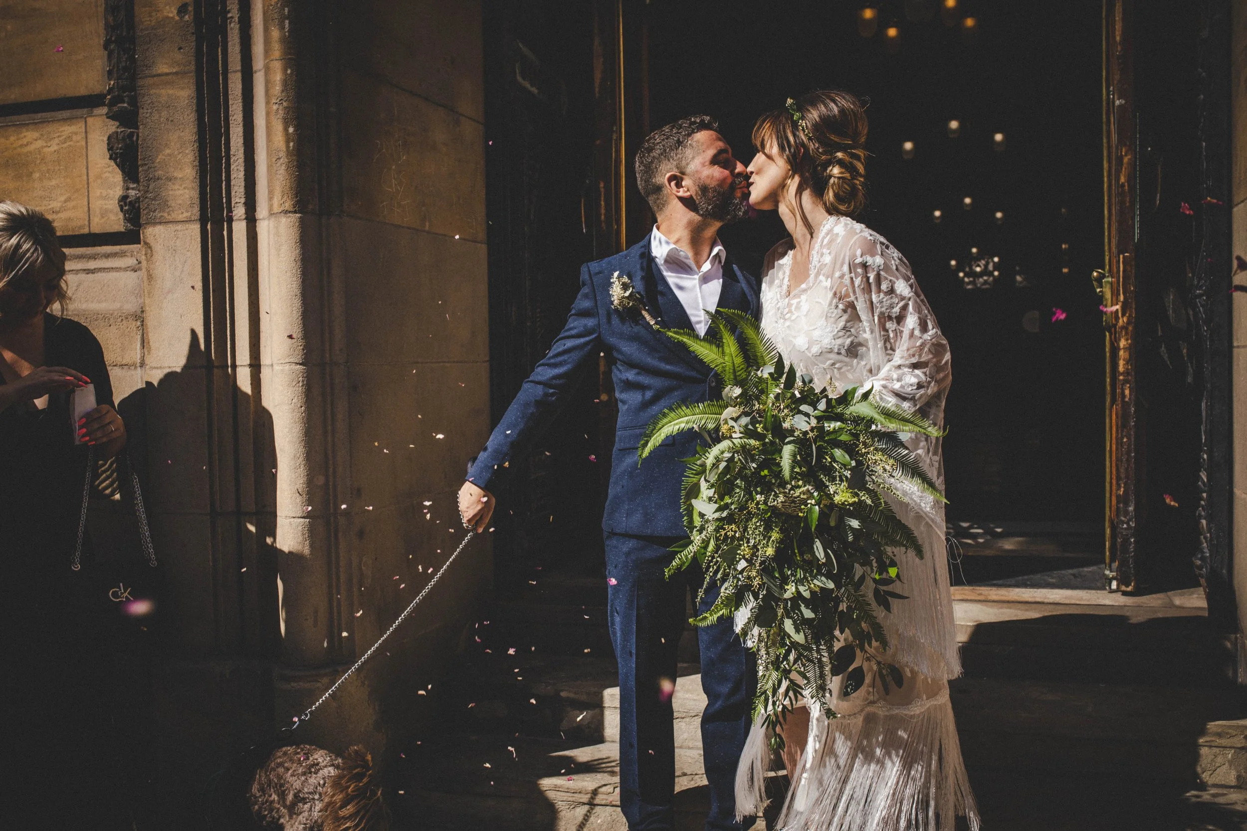A couple is sharing a kiss outside a building during a wedding ceremony, with a woman on the left taking a photo and a child at the bottom left. The groom is wearing a blue suit with a white shirt and holds a bouquet of greenery, and the bride is wea
