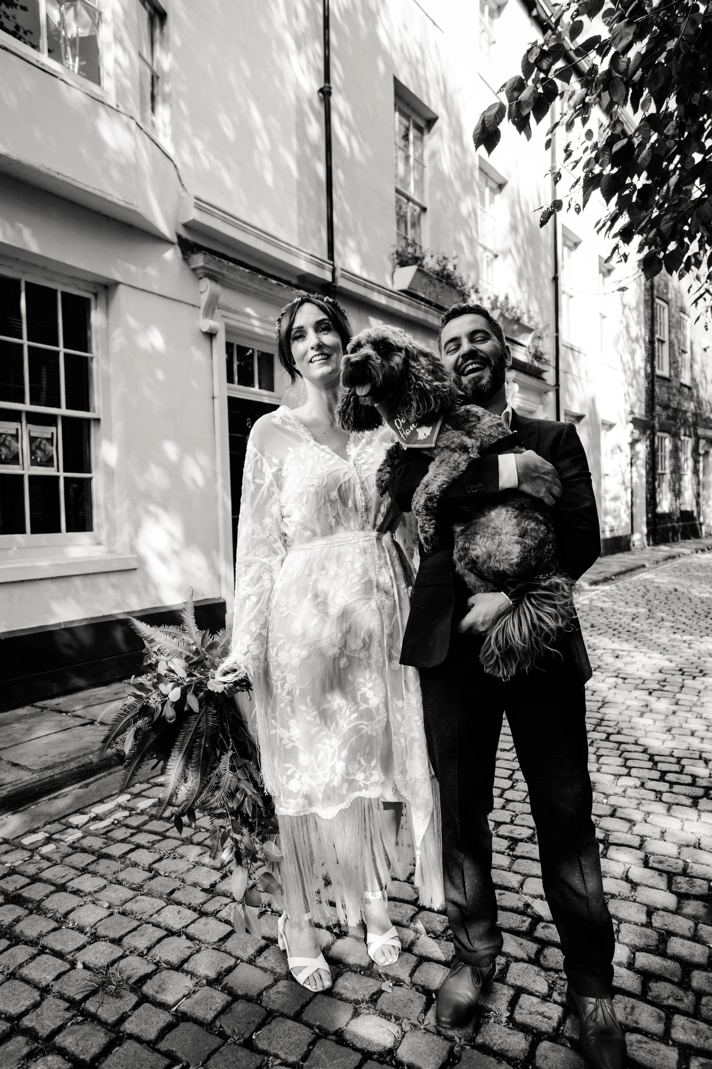 A black and white photo of a smiling bride and groom with a dog on a cobblestone street in a residential neighborhood, trees casting shadows, buildings with windows in the background.