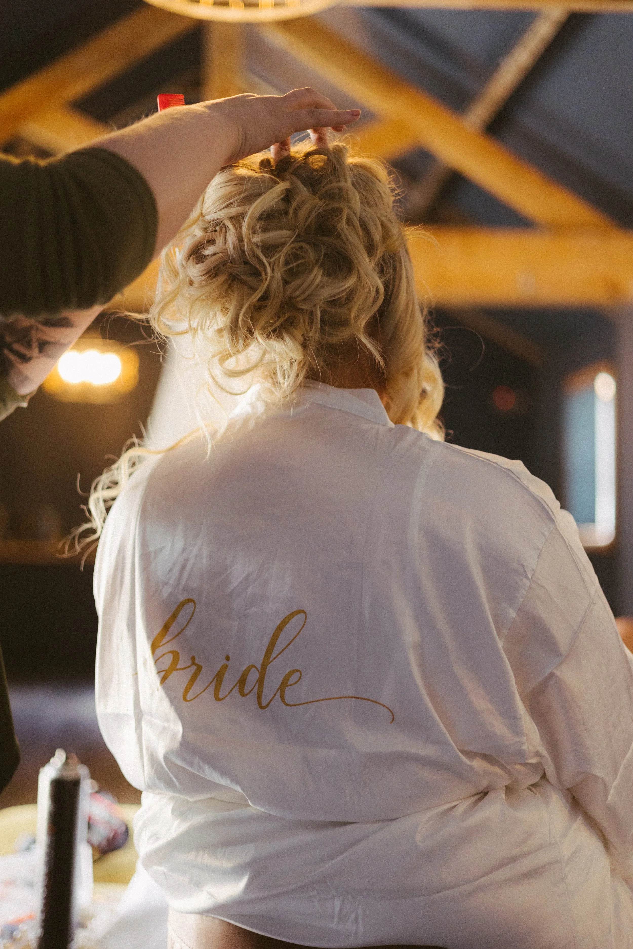 A person with curly blonde hair getting ready for their wedding, wearing a white robe with the word 'bride' written on the back, in a warmly lit room with wooden beams.