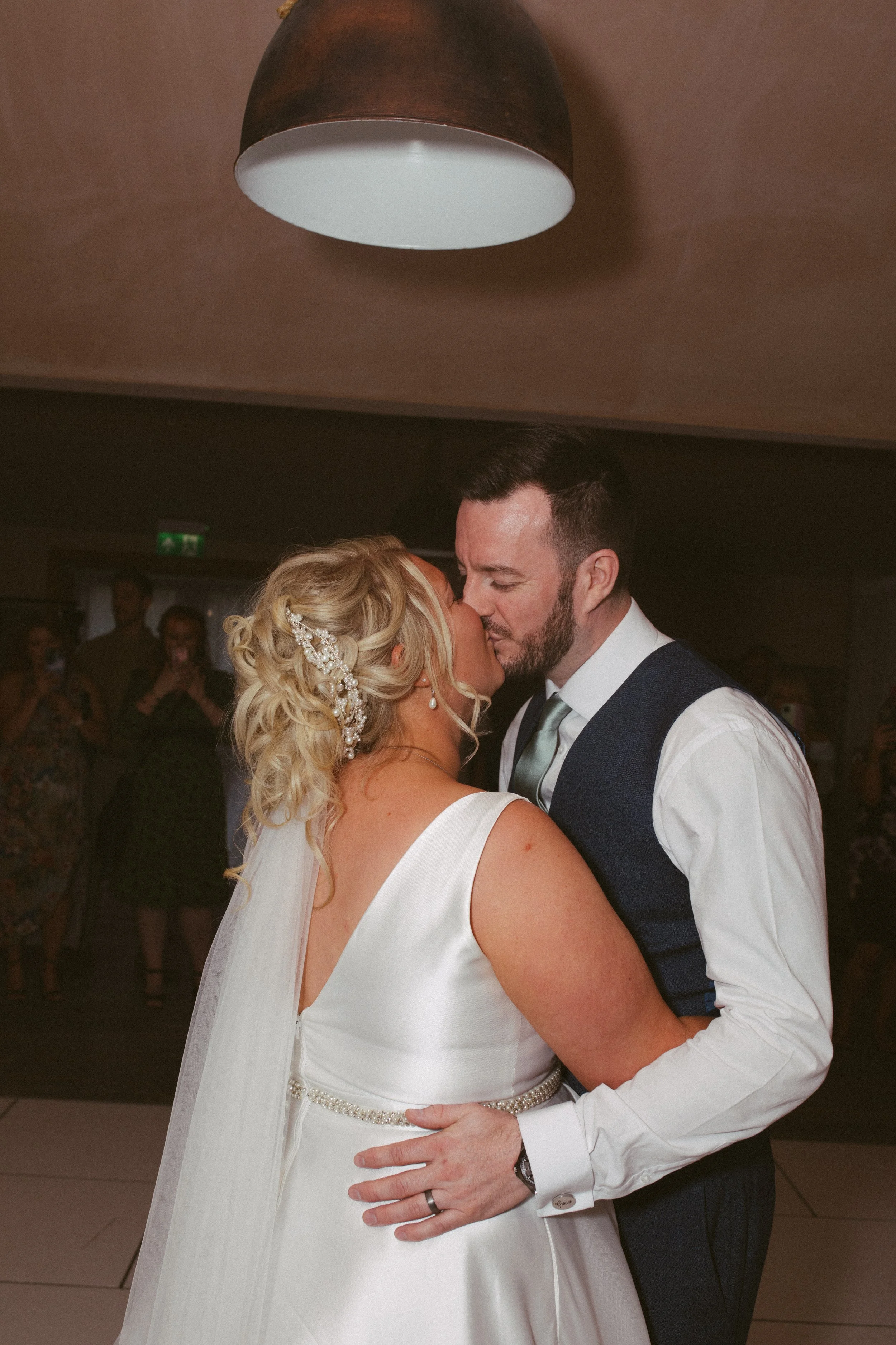 A bride and groom share a close, intimate moment on their wedding dance floor, with the bride in a white wedding dress and the groom in a vest and tie, surrounded by guests in the background.