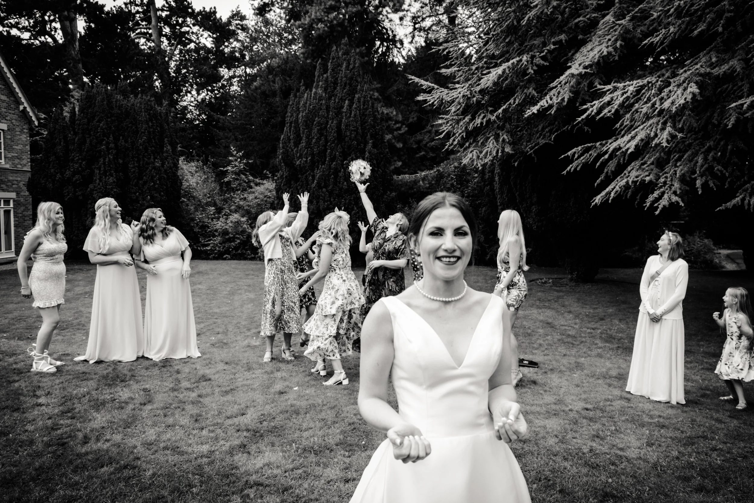 Black and white photo of women in dresses outdoors, some tossing a bouquet, smiling, with trees and a house in the background.