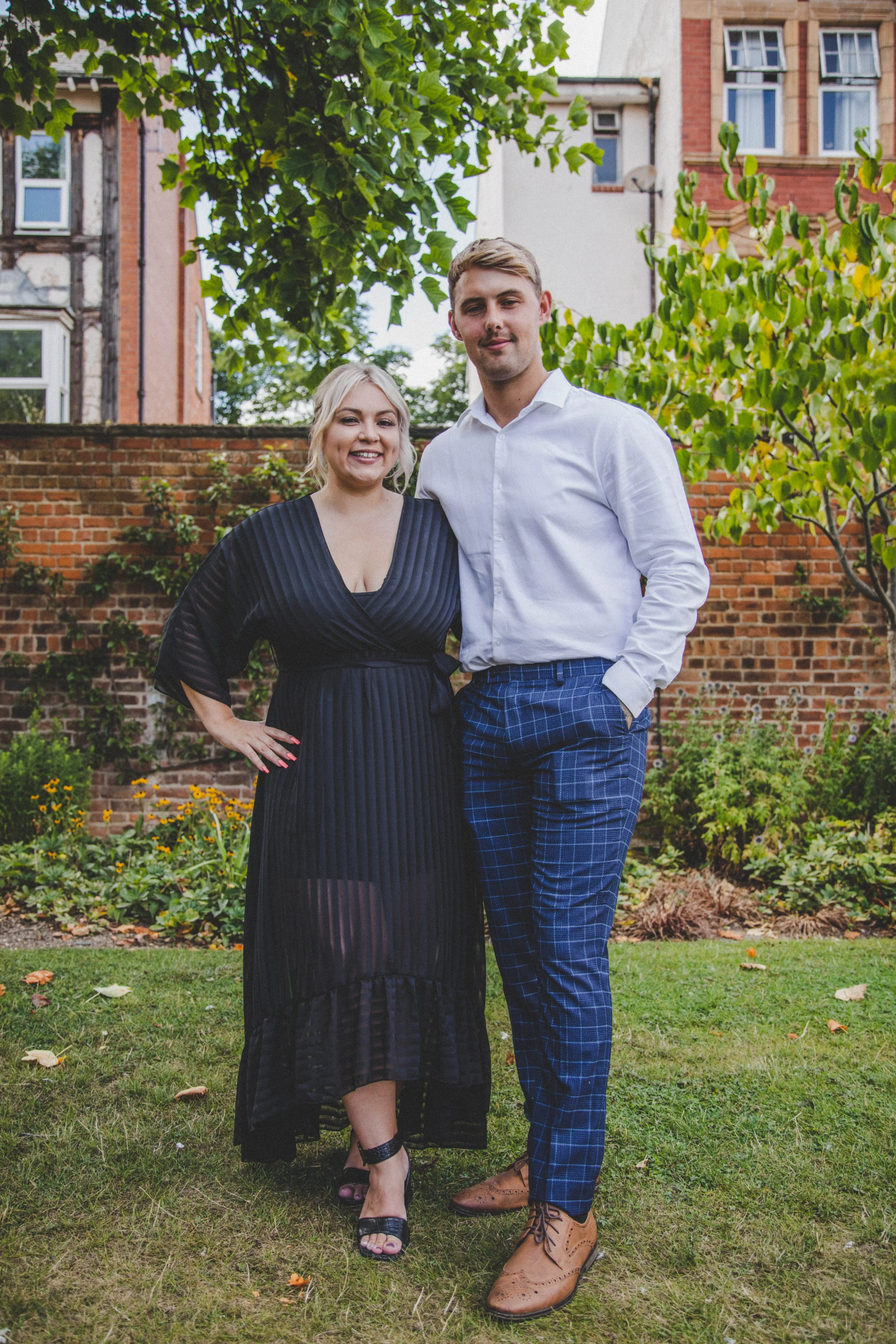 A man and woman standing together outdoors in front of a brick wall and greenery, smiling at the camera. The woman is on the left wearing a black pleated dress with black heels. The man is on the right wearing a white shirt, blue plaid pants, and bro