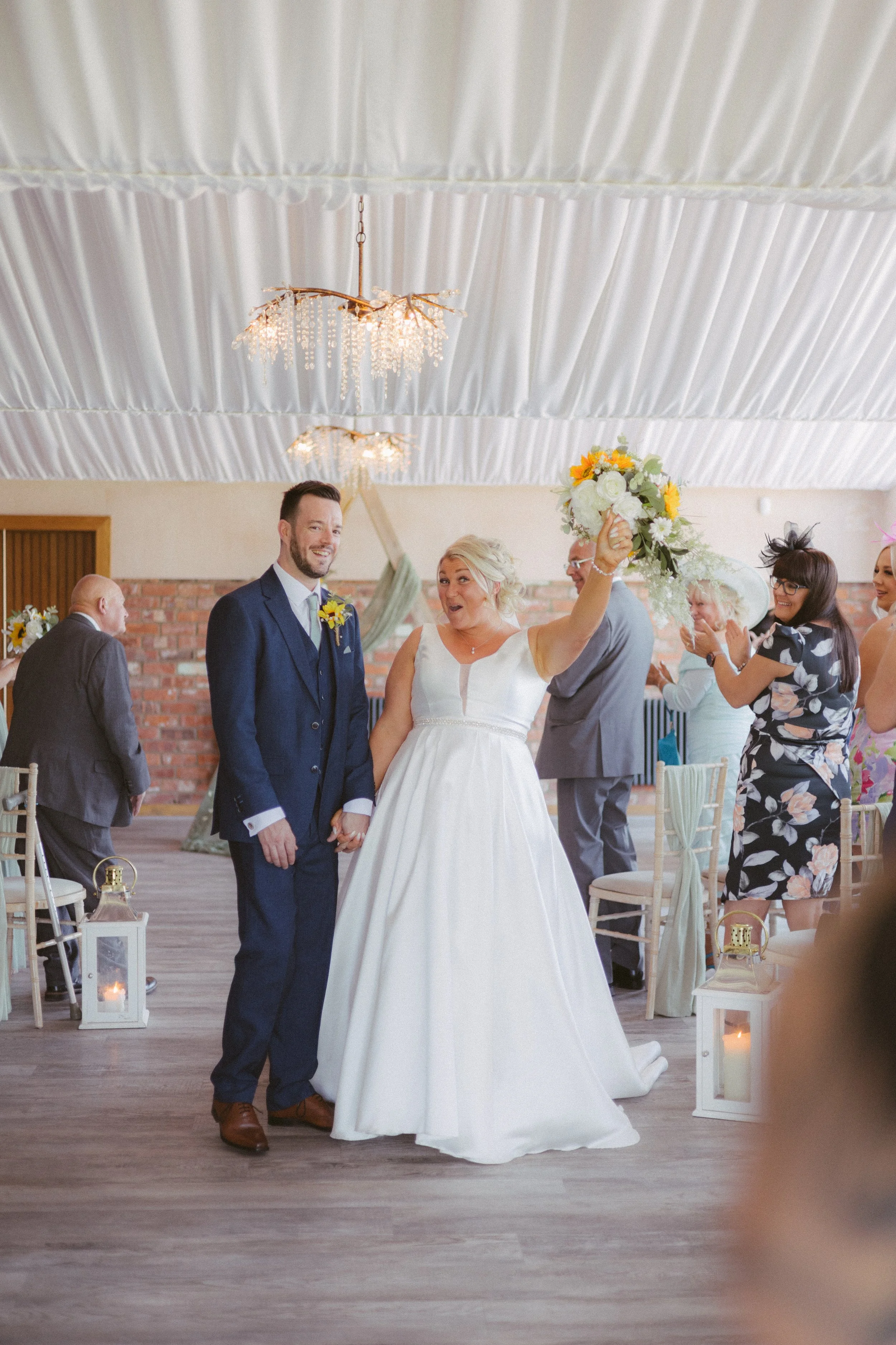 A bride and groom holding hands at their wedding ceremony, surrounded by guests in a decorated indoor venue with draped ceiling, chandeliers, and floral decorations.