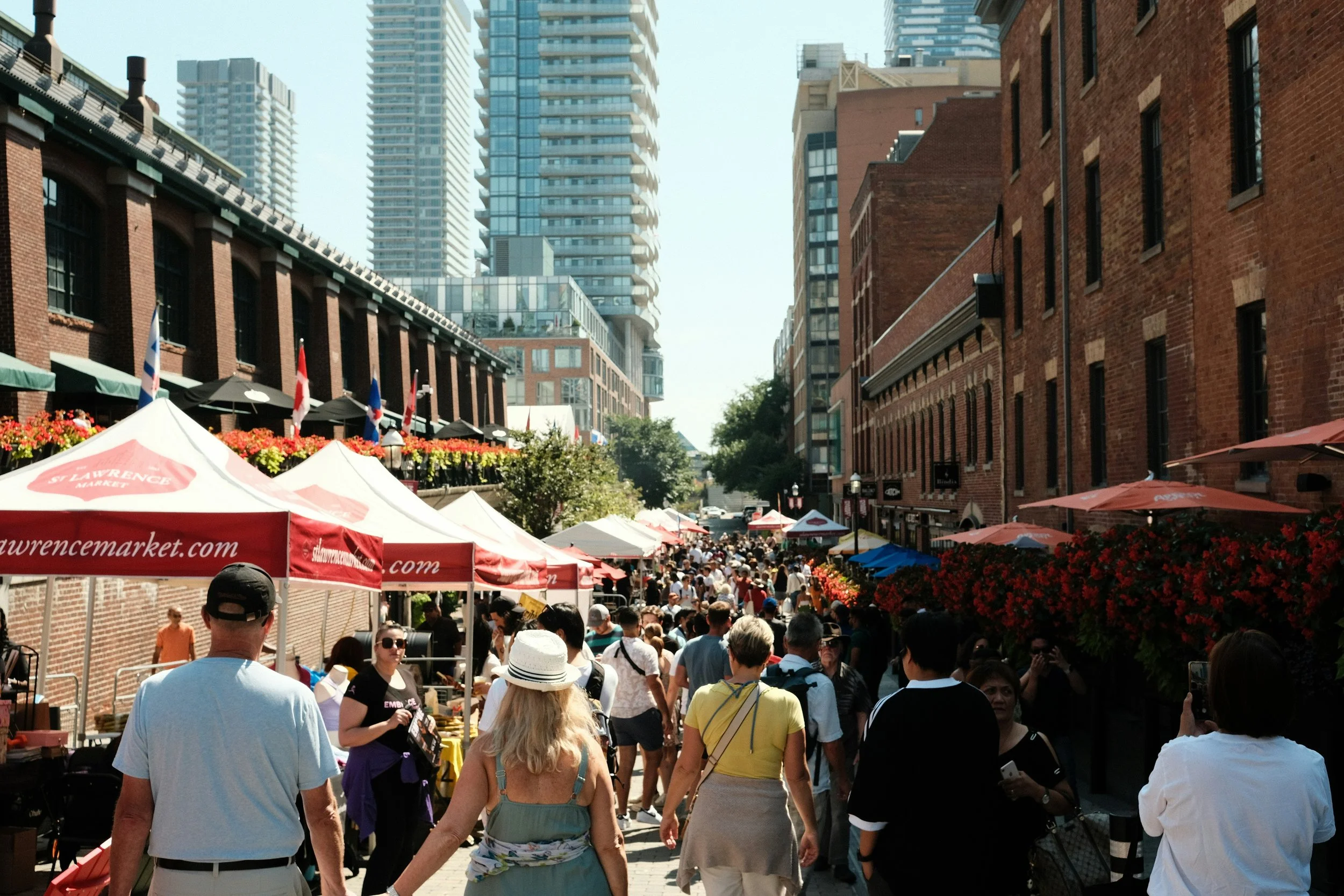 Crowded outdoor marketplace with tents, people walking, and tall buildings in the background.