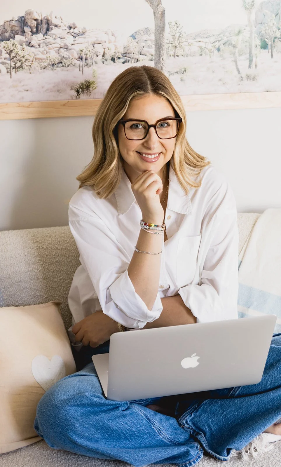 A woman with glasses and blonde hair sitting on a beige couch with a laptop on her lap, smiling and resting her chin on her hand. She is wearing a white shirt and blue jeans, with decorative bracelets on her wrist. There is a pillow with a white heart next to her, and artwork of a desert landscape with cacti and rocks in the background.