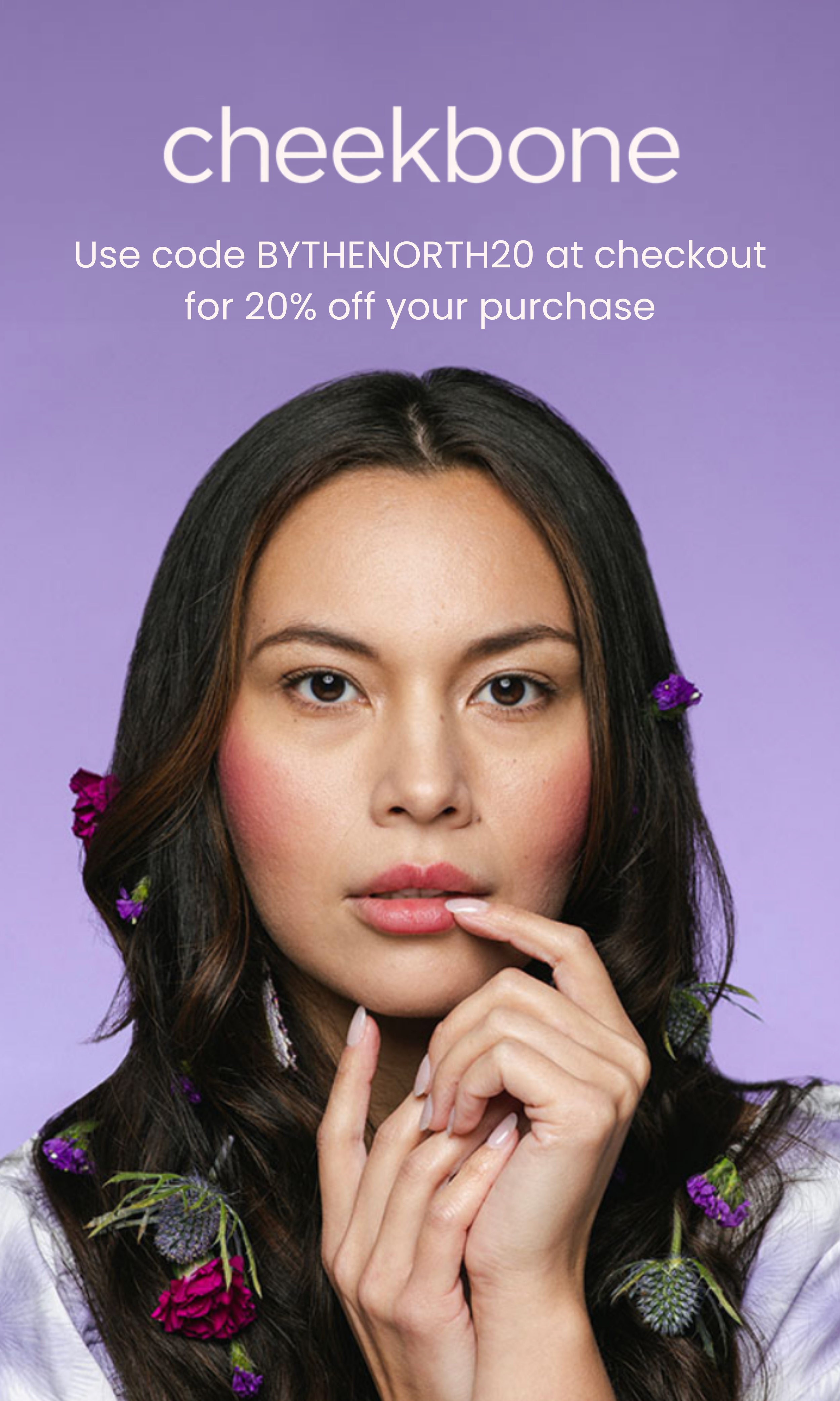 A young woman with long dark hair and flowers in her hair poses against a purple background. She touches her lips with one hand, and her expression is thoughtful.