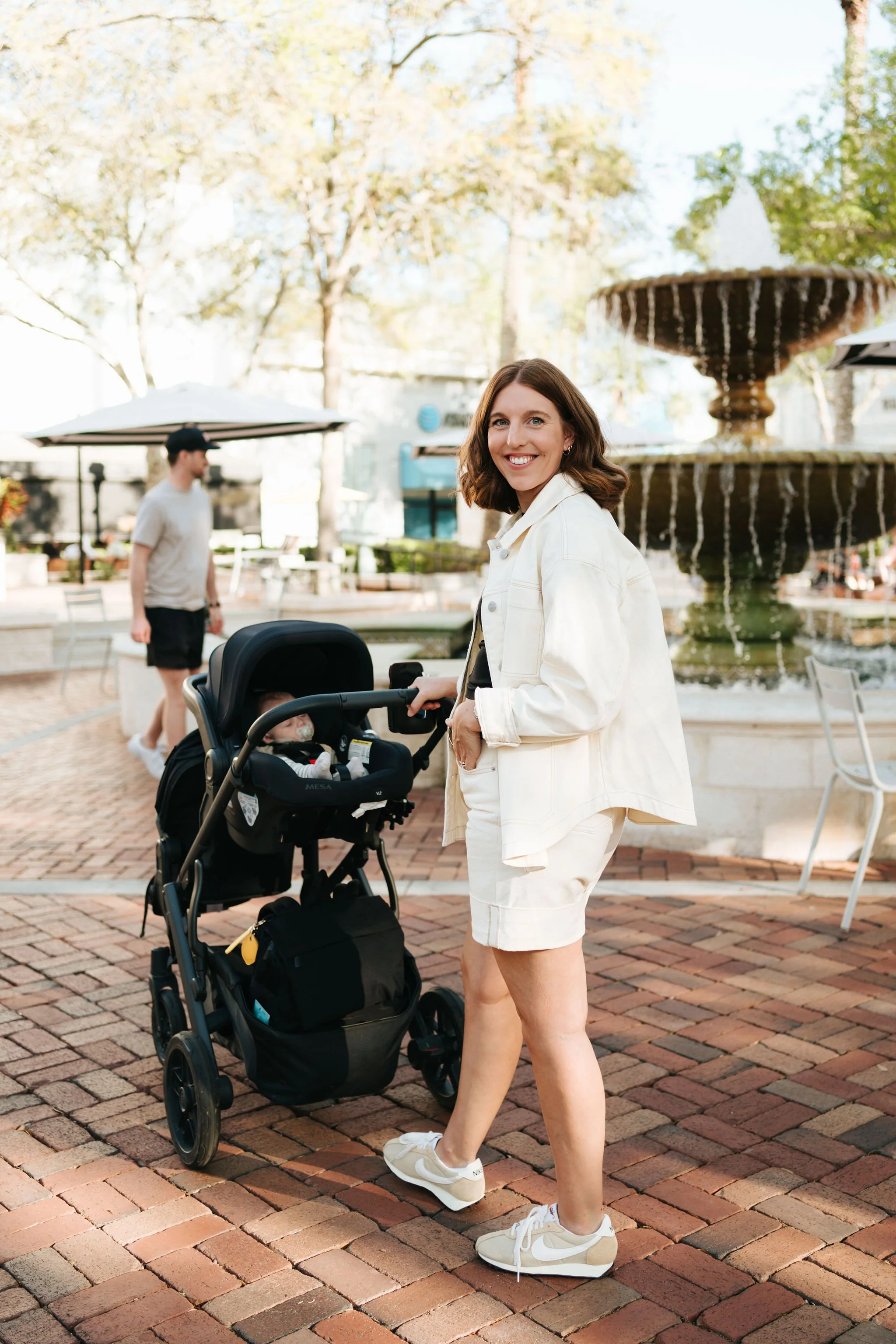 A woman with shoulder-length brown hair, dressed in an off-white denim jacket and shorts, smiling while holding a stroller with an infant inside, in an outdoor park setting near a fountain.