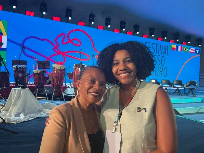 Christiane Taubira and Vanessa Vovor smiling at a conference or event with a stage in the background featuring flags and musical instruments at the Nosso Futuro Forum