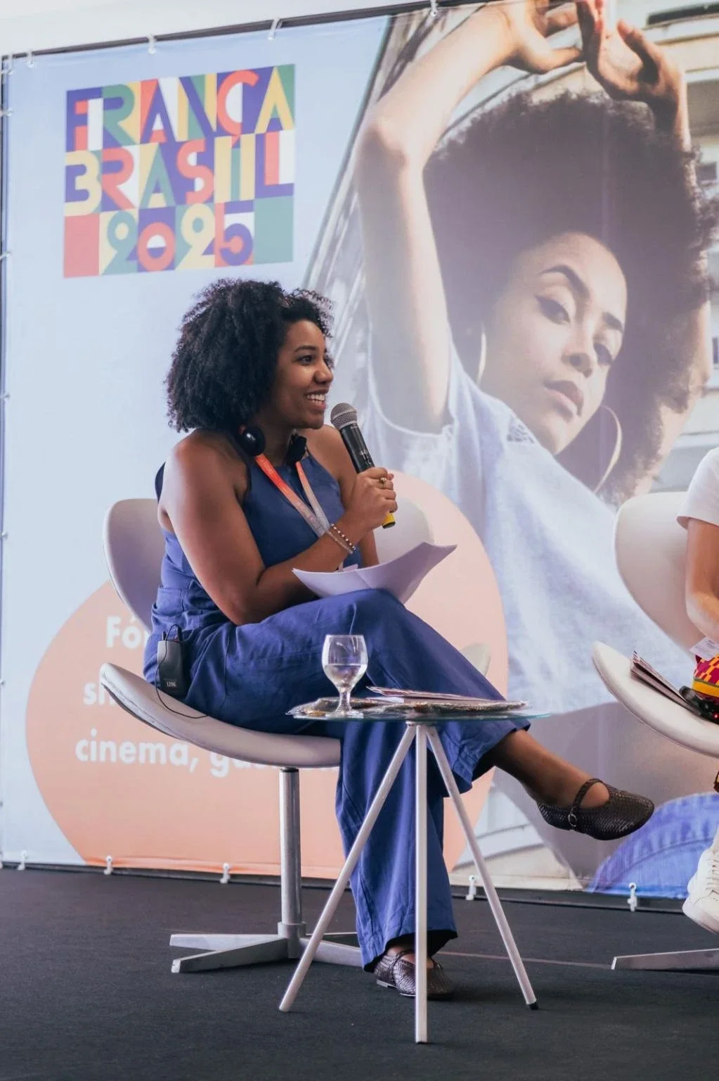 A woman with dark curly hair, wearing a blue dress, sits on a modern white chair holding a microphone and some papers. She is smiling during a panel discussion at an event called 'France Brasil 2023.' A large poster behind her features a woman with voluminous hair and the event's colorful logo.