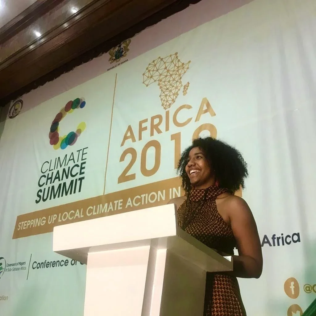 A woman is standing at a podium, smiling, in front of a large banner for the Climate Chance Africa 2019 Summit, with the theme "Stepping Up Local Climate Action in Africa."