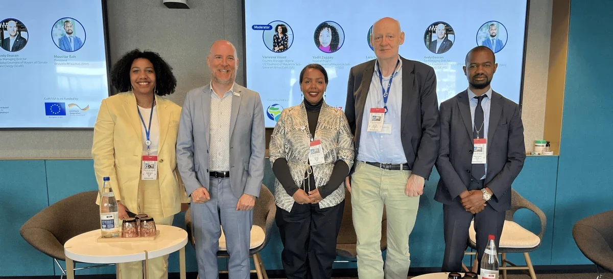 Five diverse professionals standing side by side in a conference room, smiling at the camera, with large screens displaying names and titles in the background.