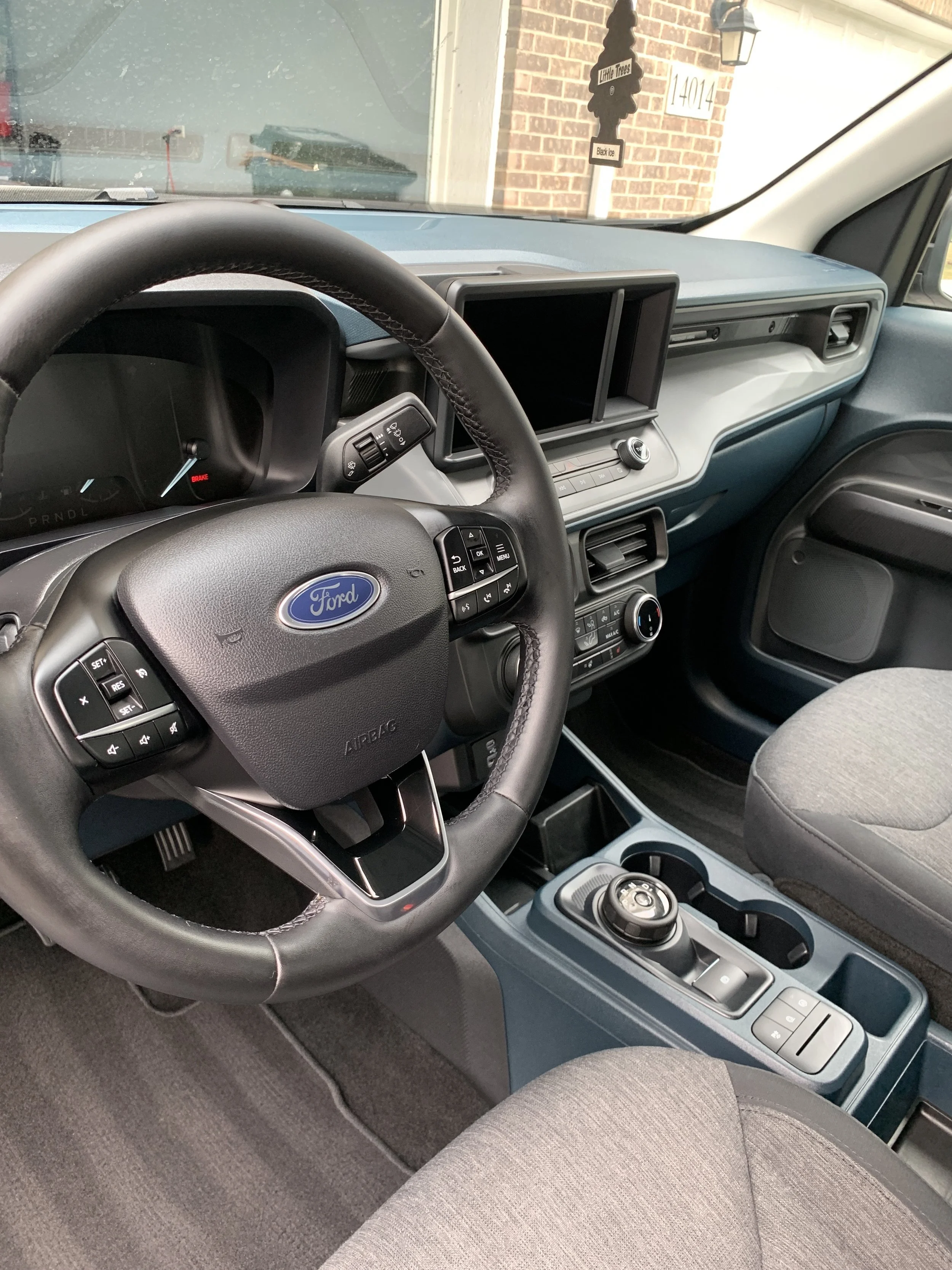 Interior of a Ford vehicle showing the steering wheel, dashboard, gear shifter, and front seats with an empty display screen and brick house visible through the windshield.