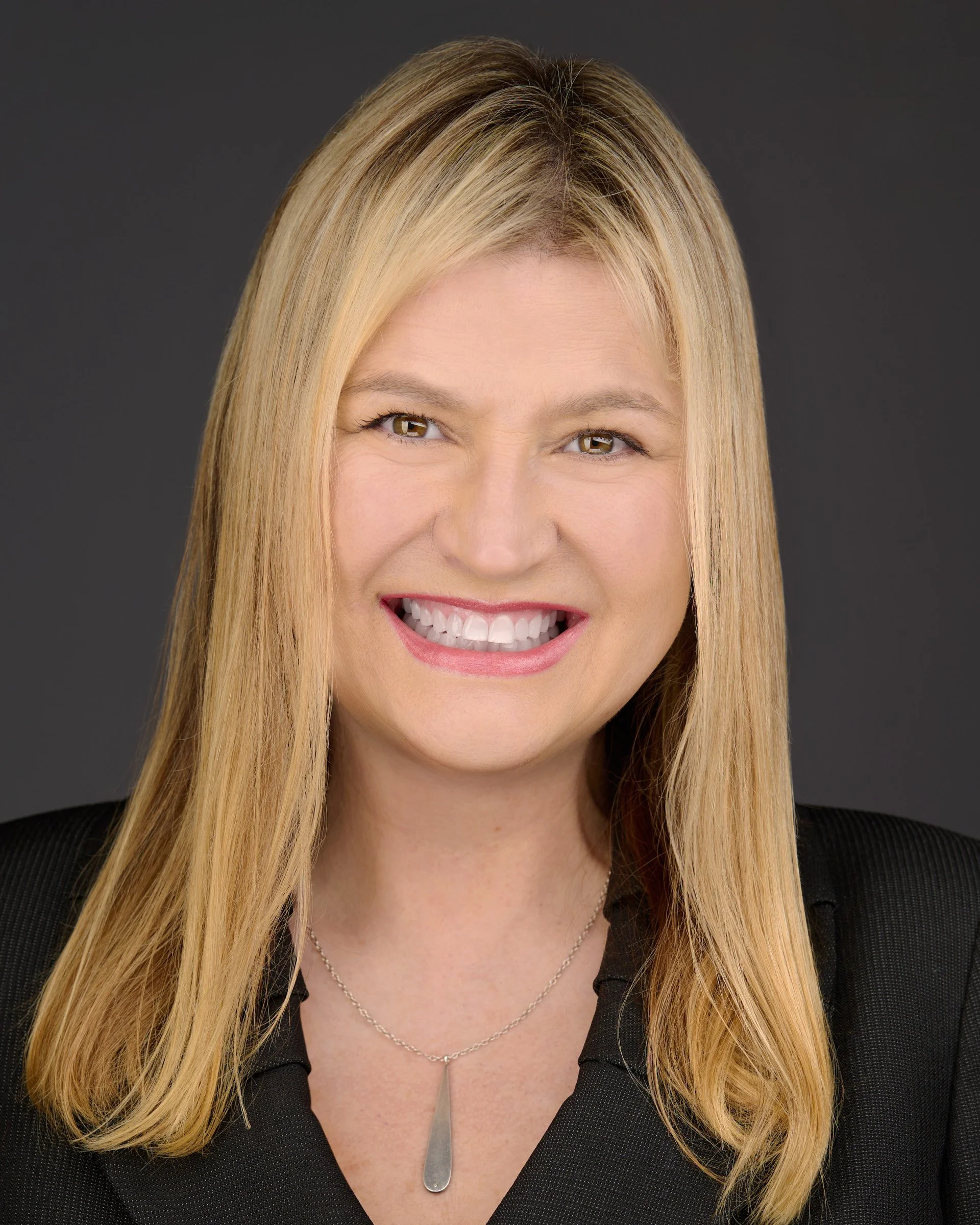 Headshot of a smiling woman with long blonde hair wearing a black top and a silver necklace with a pendant