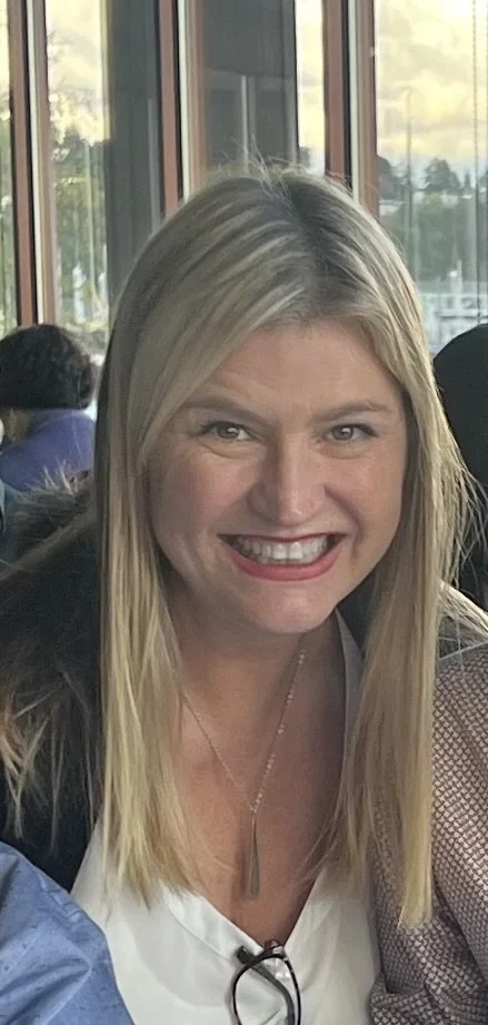 A woman smiling with long blonde hair, wearing a white top and a necklace, sitting indoors near large windows.