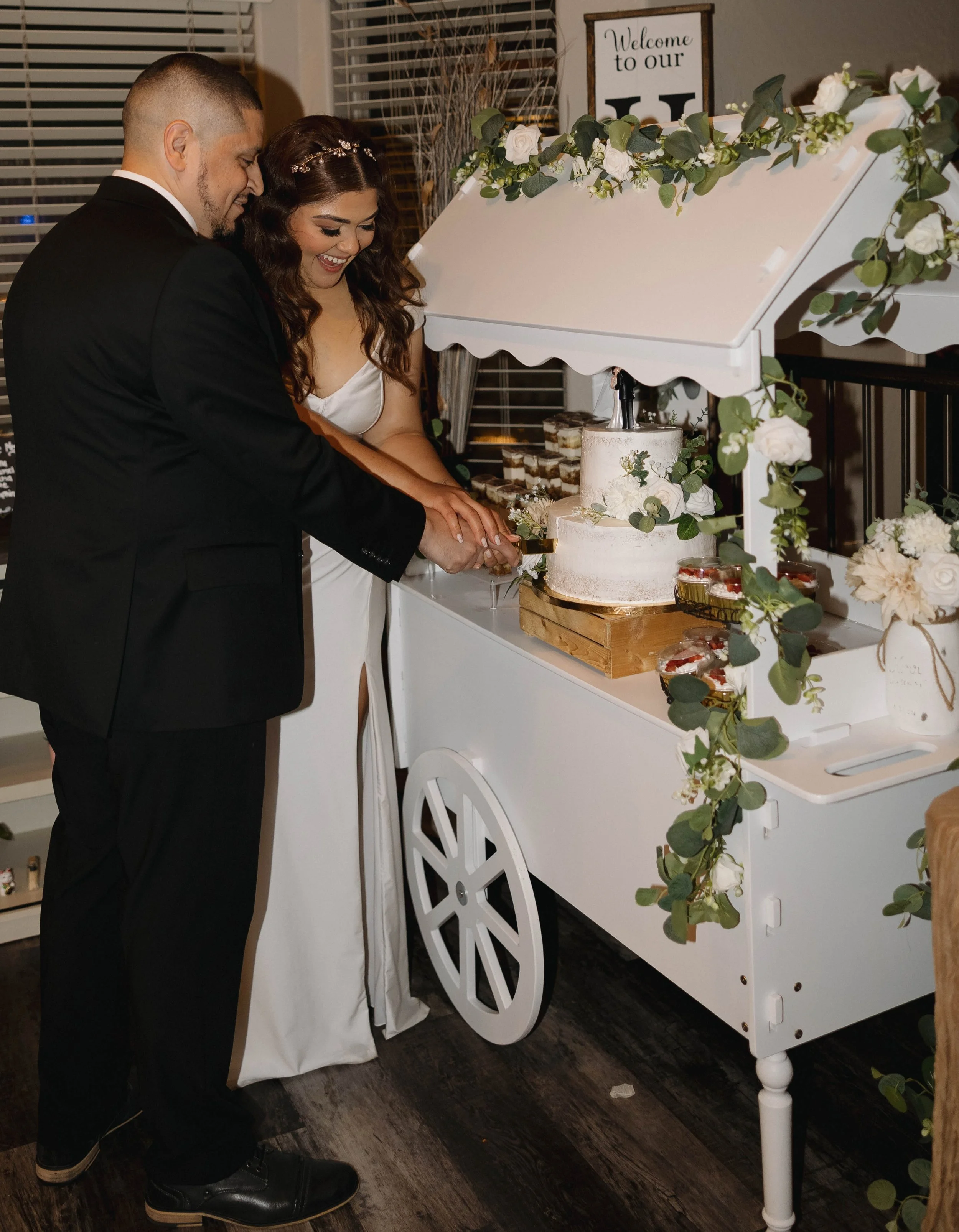 A bride and groom cut a wedding cake together at their reception, with a floral and greenery decoration on a white cart.