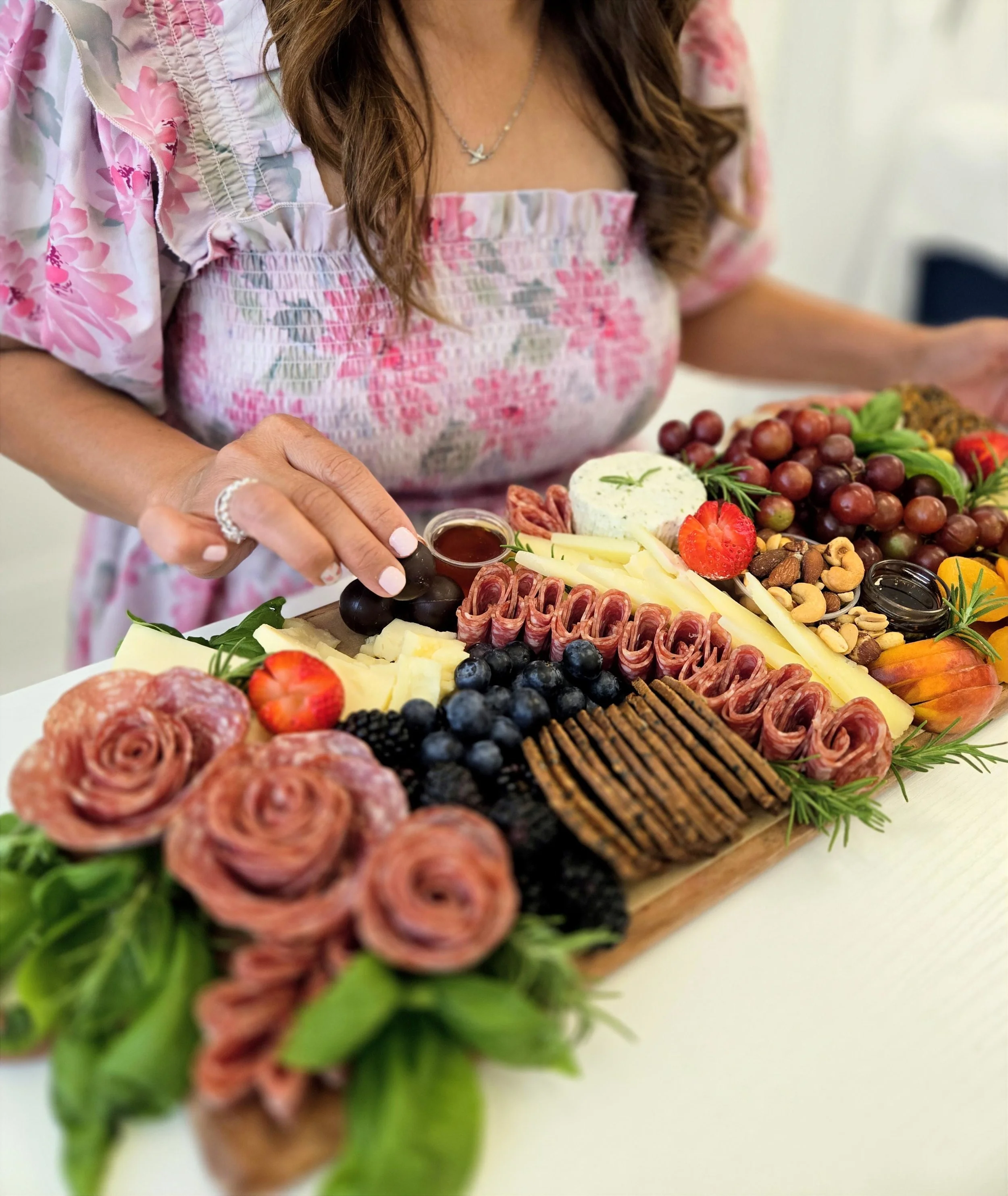 Close-up of a woman in a floral dress preparing a charcuterie board with cheese, grapes, strawberries, nuts, and cured meats, garnished with herbs, on a white surface.