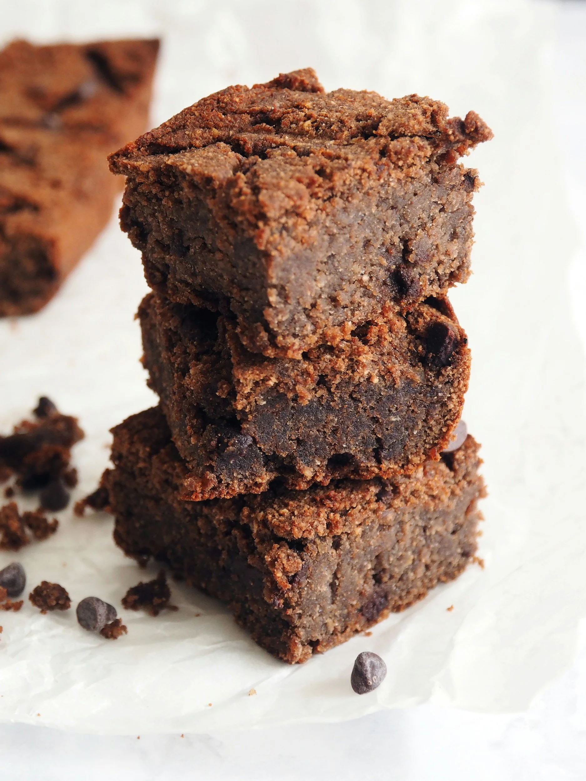 Three stacked chocolate brownies with a crumbly top, on white parchment paper, with chocolate chips scattered around.