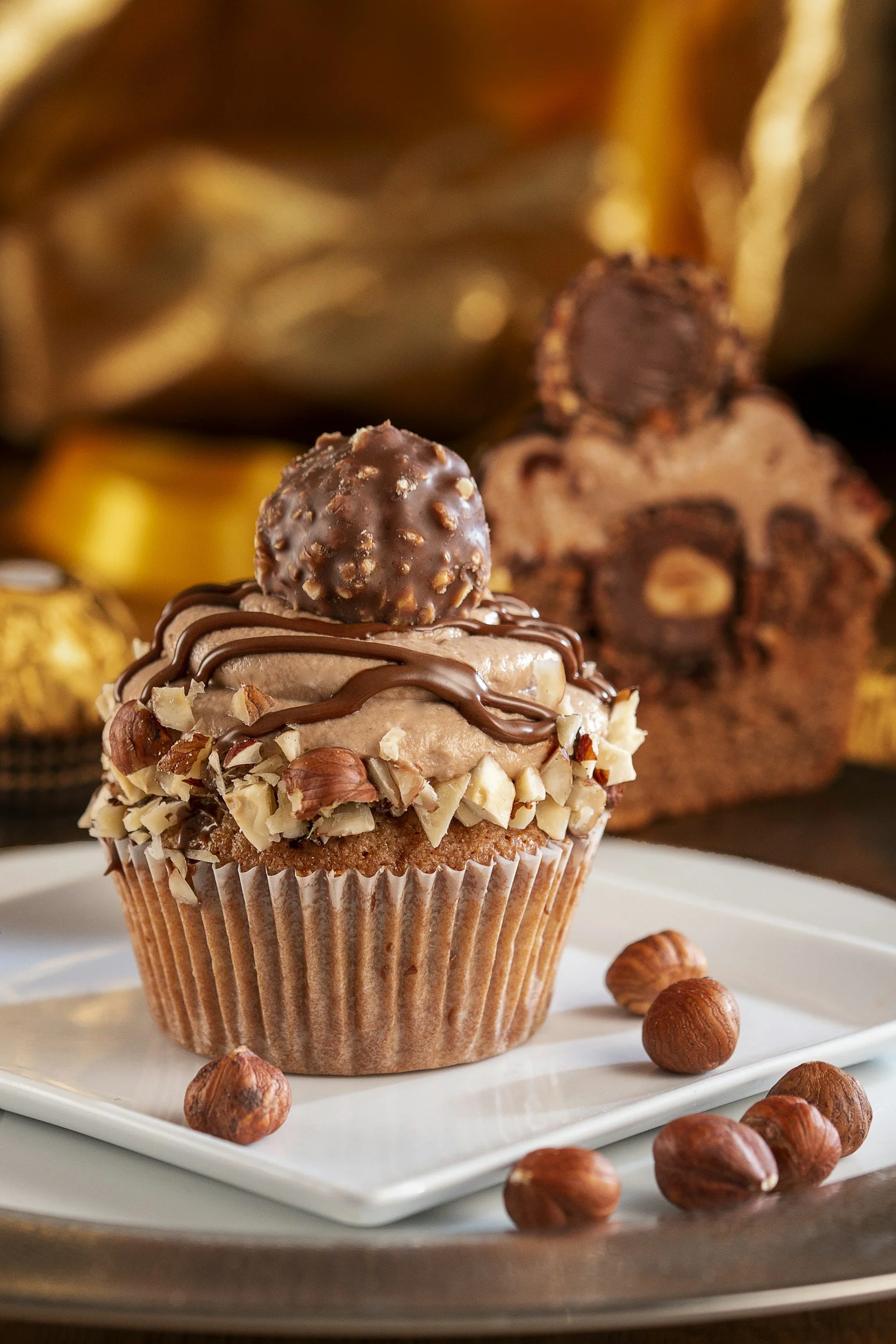 Chocolate cupcake topped with a chocolate truffle, chopped nuts, and drizzled with chocolate, placed on a white square plate with hazelnuts around it.