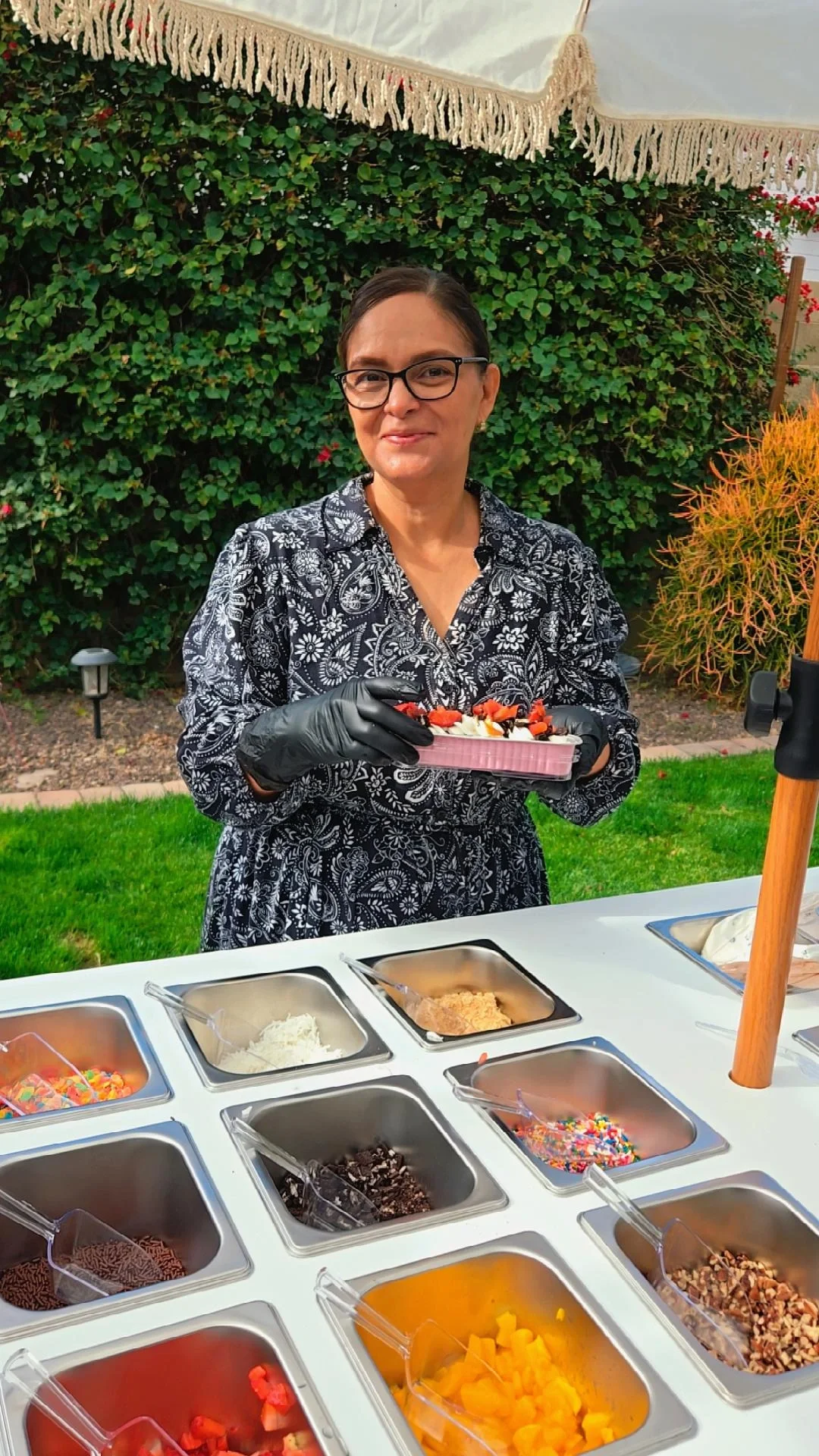 Woman serving frozen yogurt at an outdoor station with various toppings, wearing black gloves and glasses, in a backyard with green bushes and a white canopy overhead.