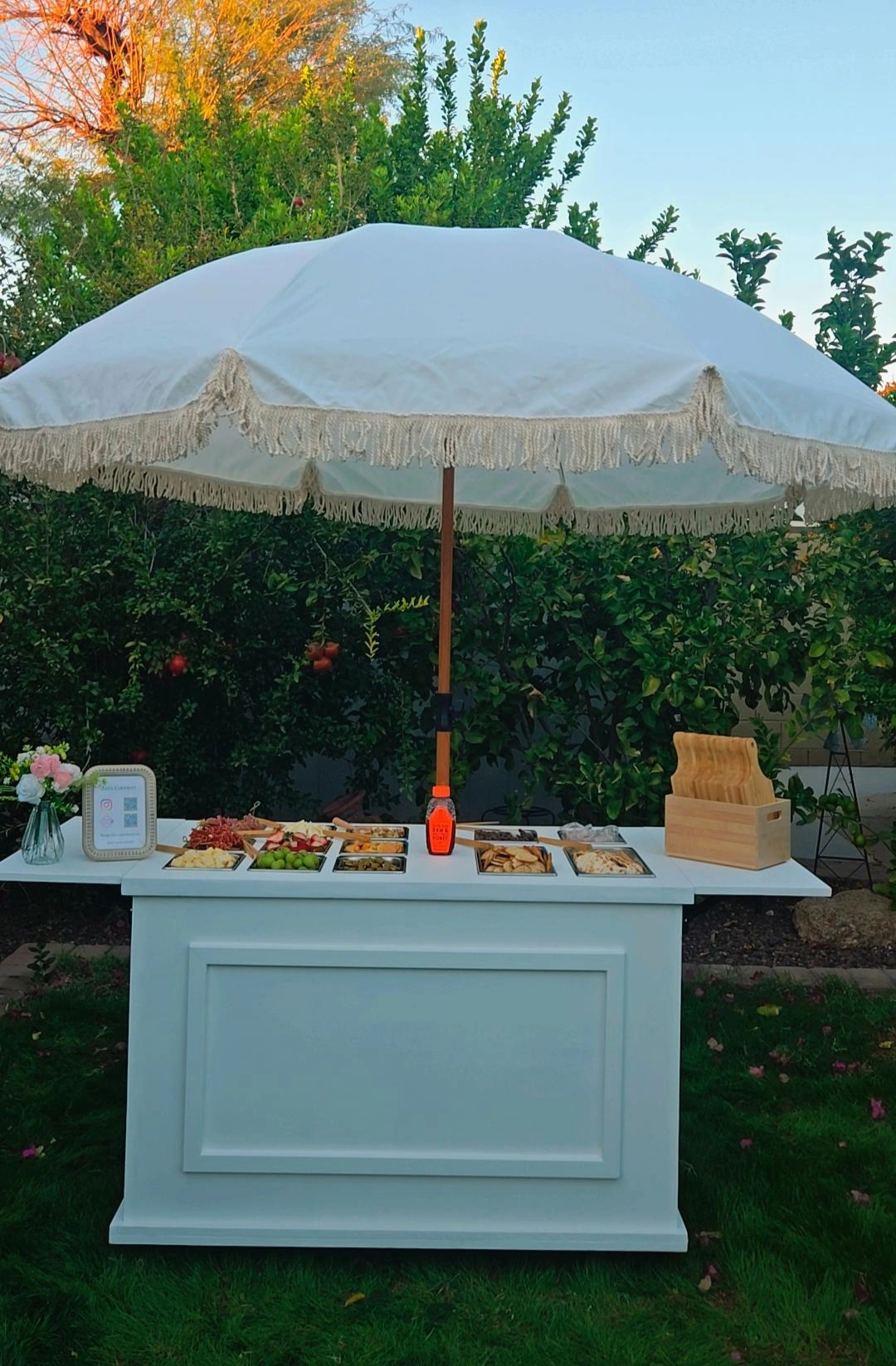 Outdoor food table under a large white umbrella with fringe, holding various snacks, fruits, and condiments, set in a garden with trees and grass.