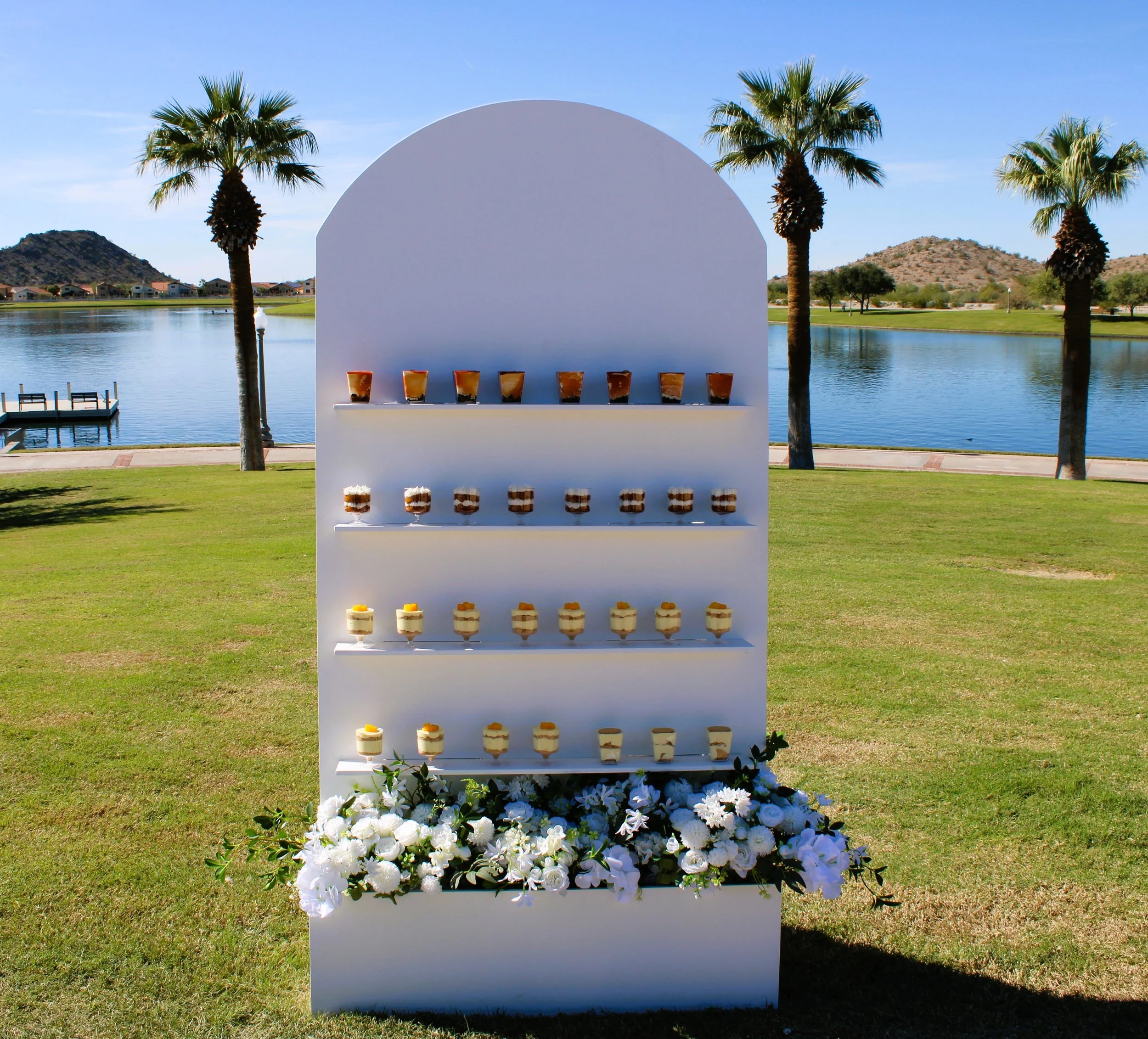 A white display stand with multiple tiers of desserts, decorated with white flowers, set in a grassy area near a lake with palm trees and mountains in the background perfect for weddings and showers.