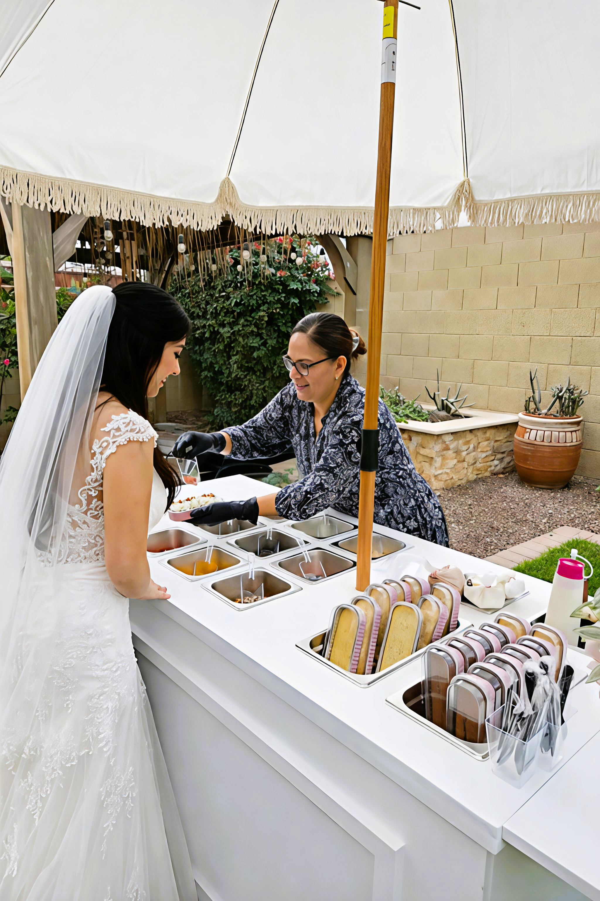 Bride at her wedding reception being served from a dessert or candy station under a white umbrella, with a woman in glasses helping her.