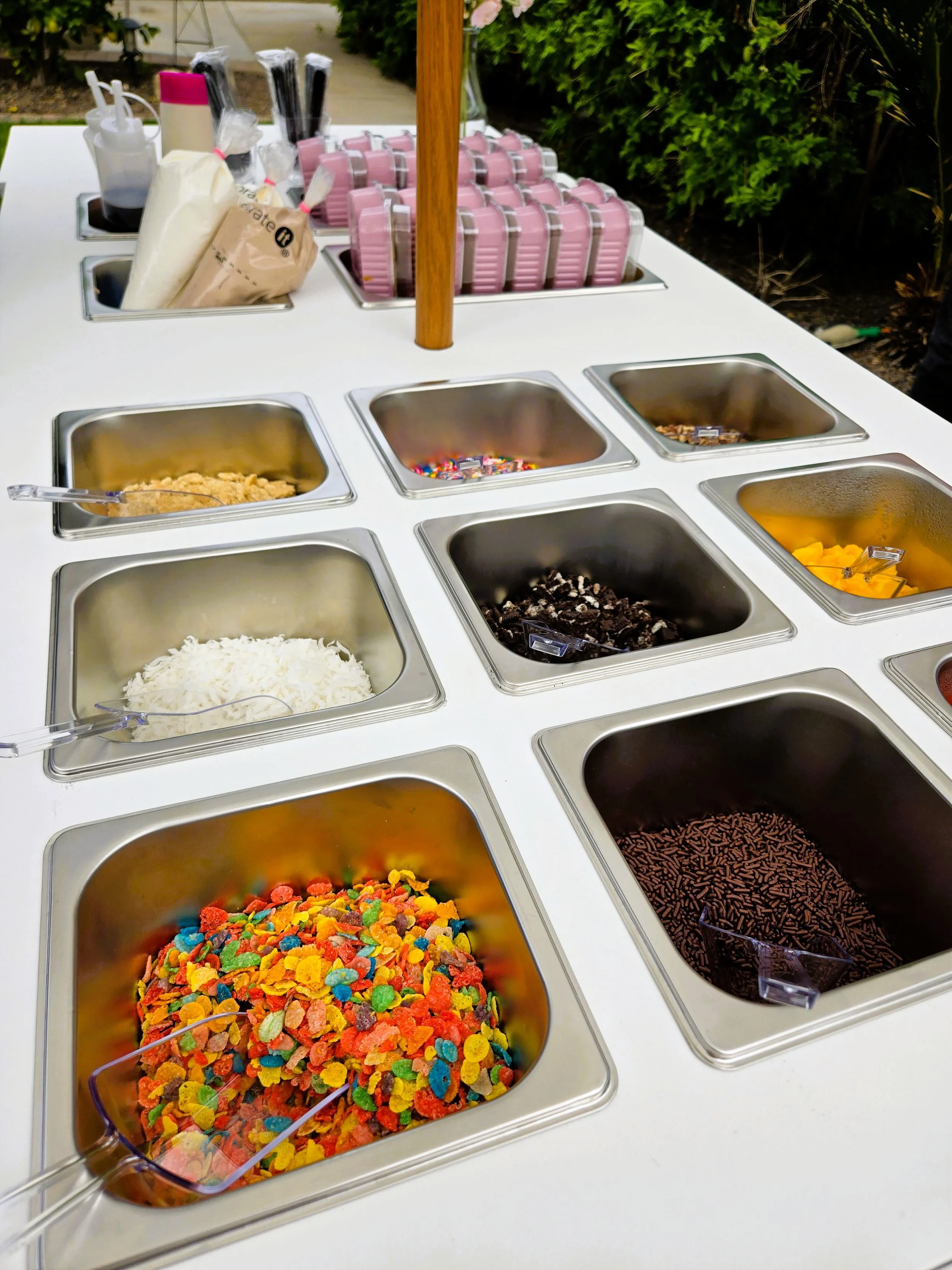 Ice cream toppings at a self-serve station, including colorful cereal, sprinkles, crushed cookies, and syrups.