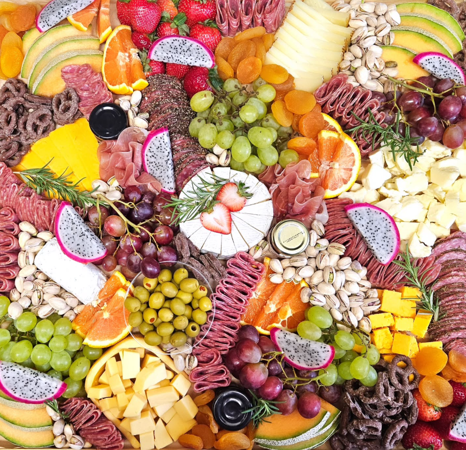 A colorful charcuterie board with assorted cheeses, fruits, nuts, dried fruits, salami, crackers, and herbs.