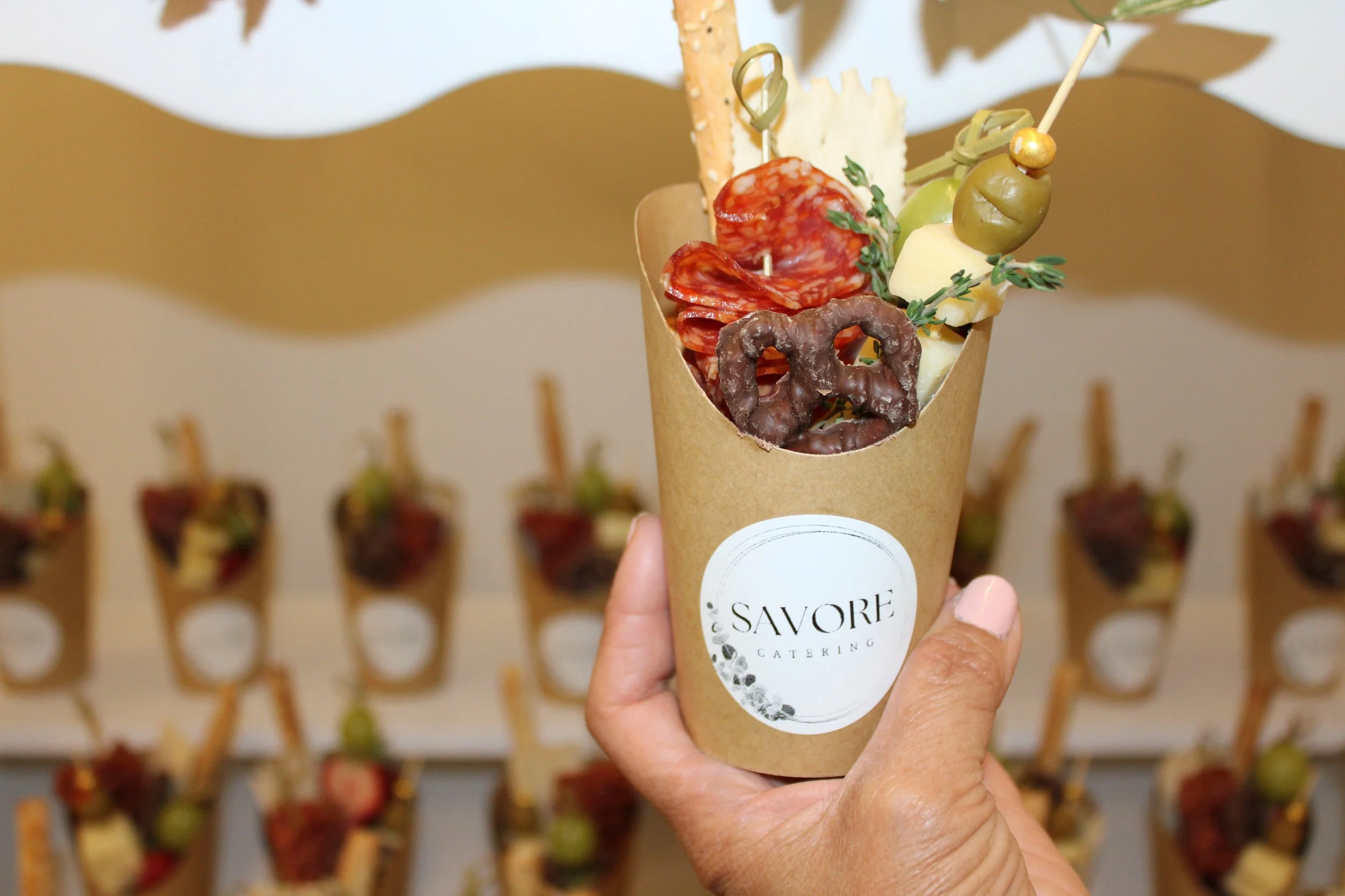 Hand holding a cone with assorted charcuterie items including cured meats, cheese, olives, and breadsticks, with a background of similar cones displayed on a wall.