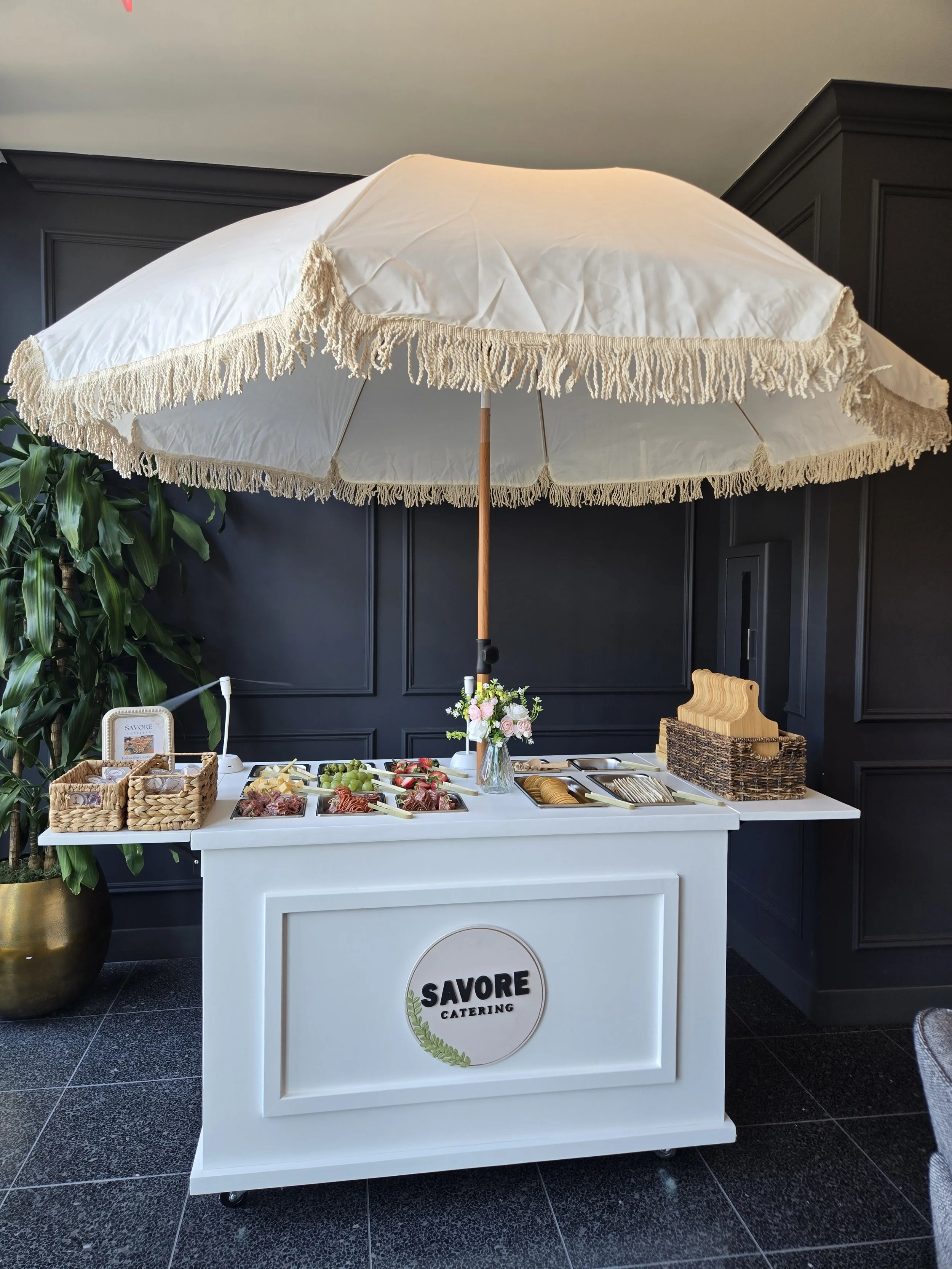 Catering station with a white table, a large white parasol with beige fringes, and a dark wall behind. The station has fresh fruits, snacks, and flowers, with a sign that reads "SAVORE Catering." There is a large plant to the left.