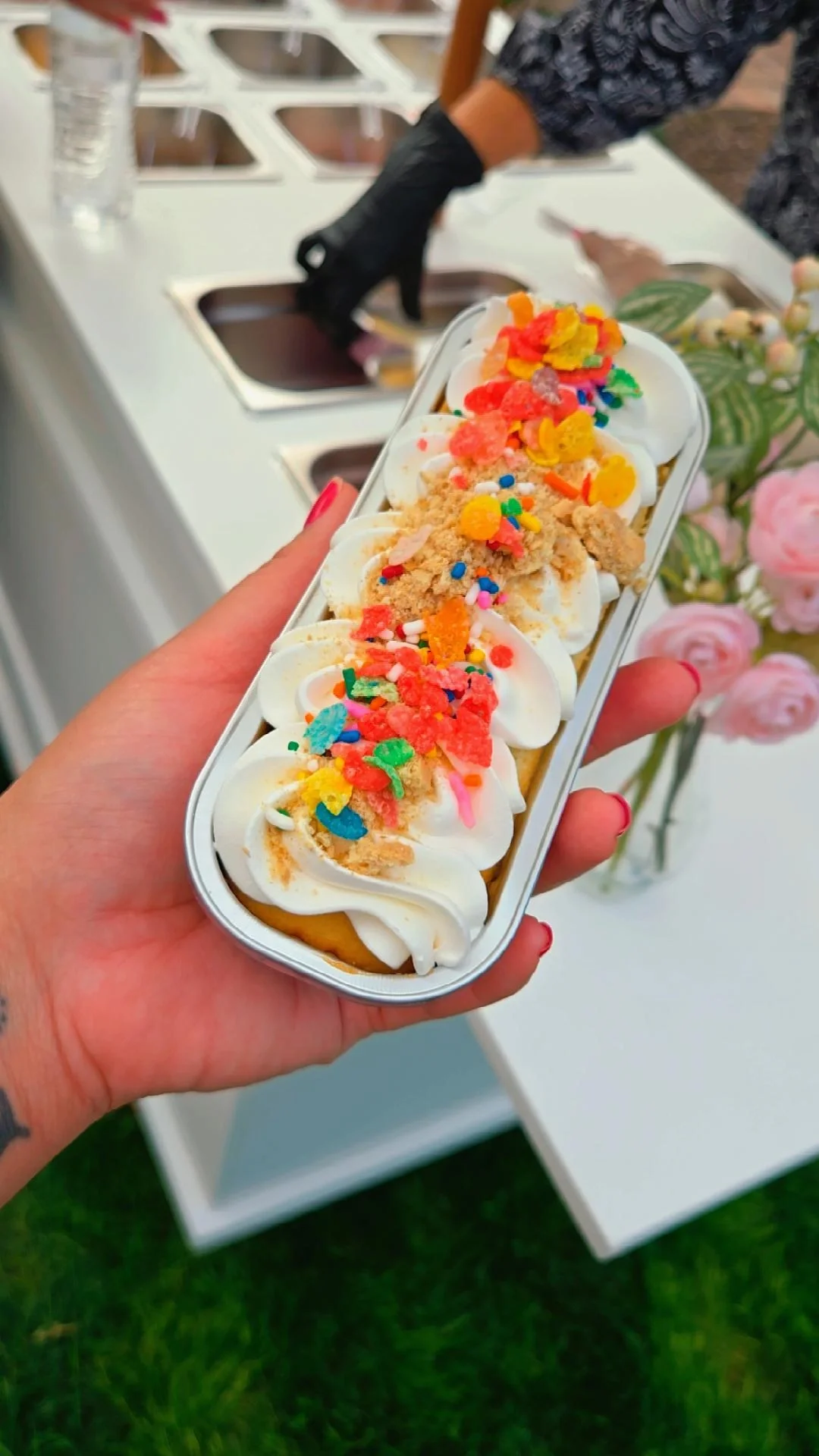 Close-up of a hand holding a decorated ice cream snack with whipped cream, colorful sprinkles, crushed cookies, and fruity toppings, with a food counter and people in the background.