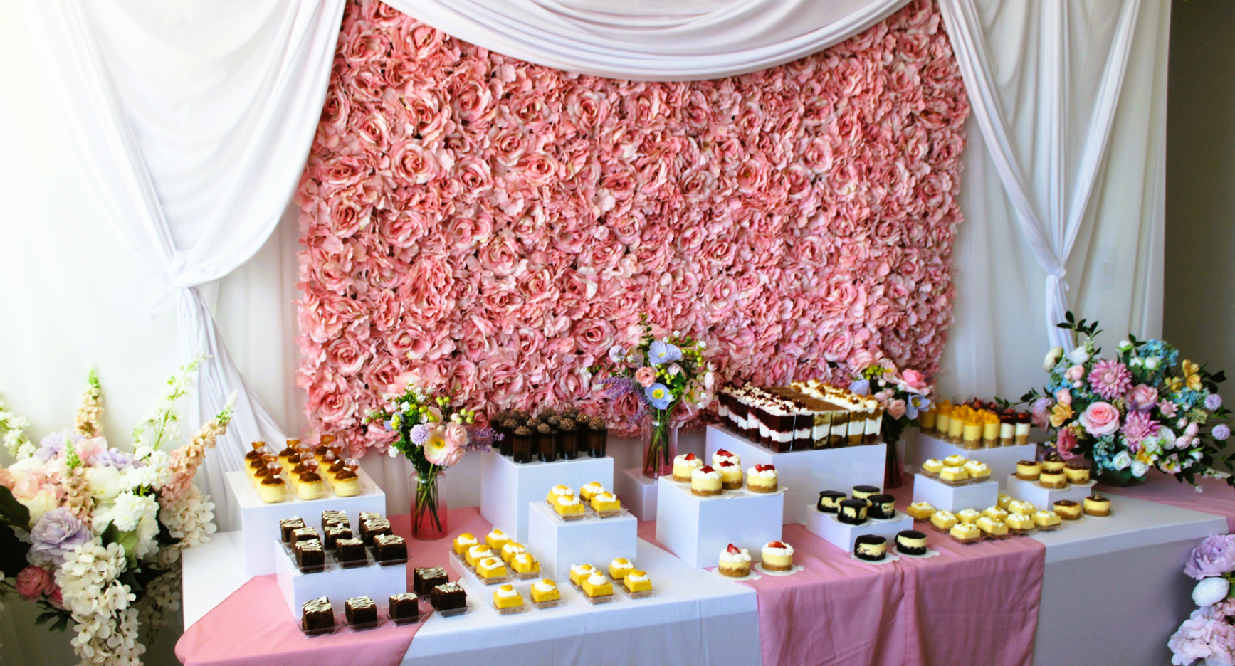 Dessert table with various small cakes and sweets, decorated with large flower arrangements, against a pink floral backdrop with white drapes.
