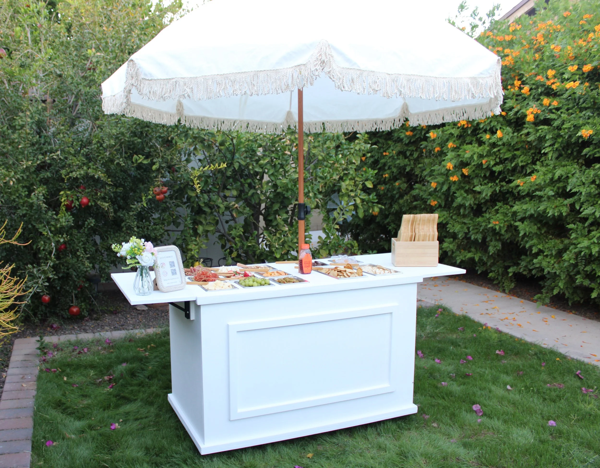 A white outdoor table with a canopy, set up for a gathering, with various foods and a flower arrangement, in a garden with trees and bushes with orange flowers.