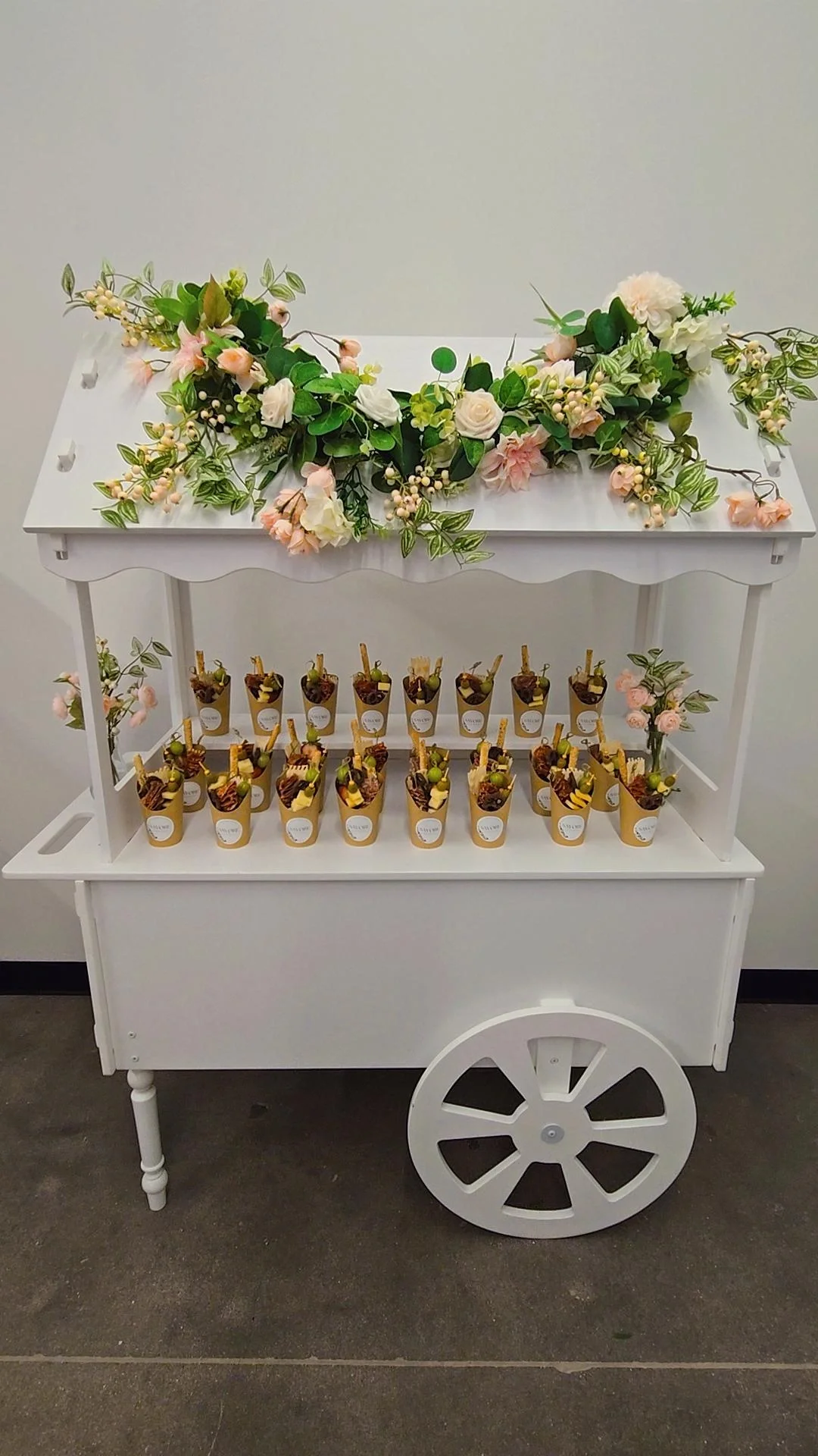 White dessert cart with a floral display on top and small dessert cups lined on the middle shelf.