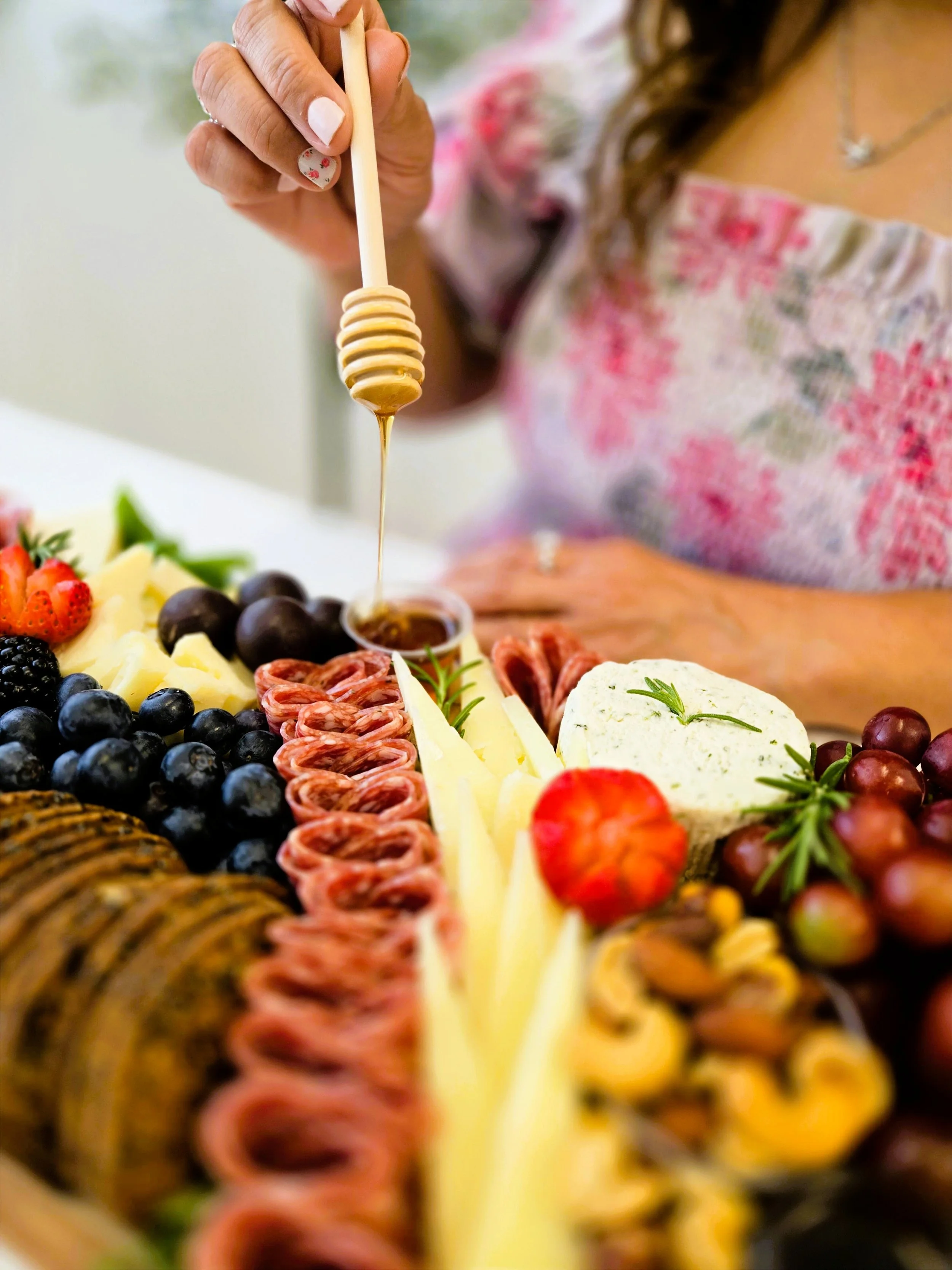 A person drizzling honey onto a cheese and fruit platter that includes grapes, strawberries, blueberries, and slices of cheese.